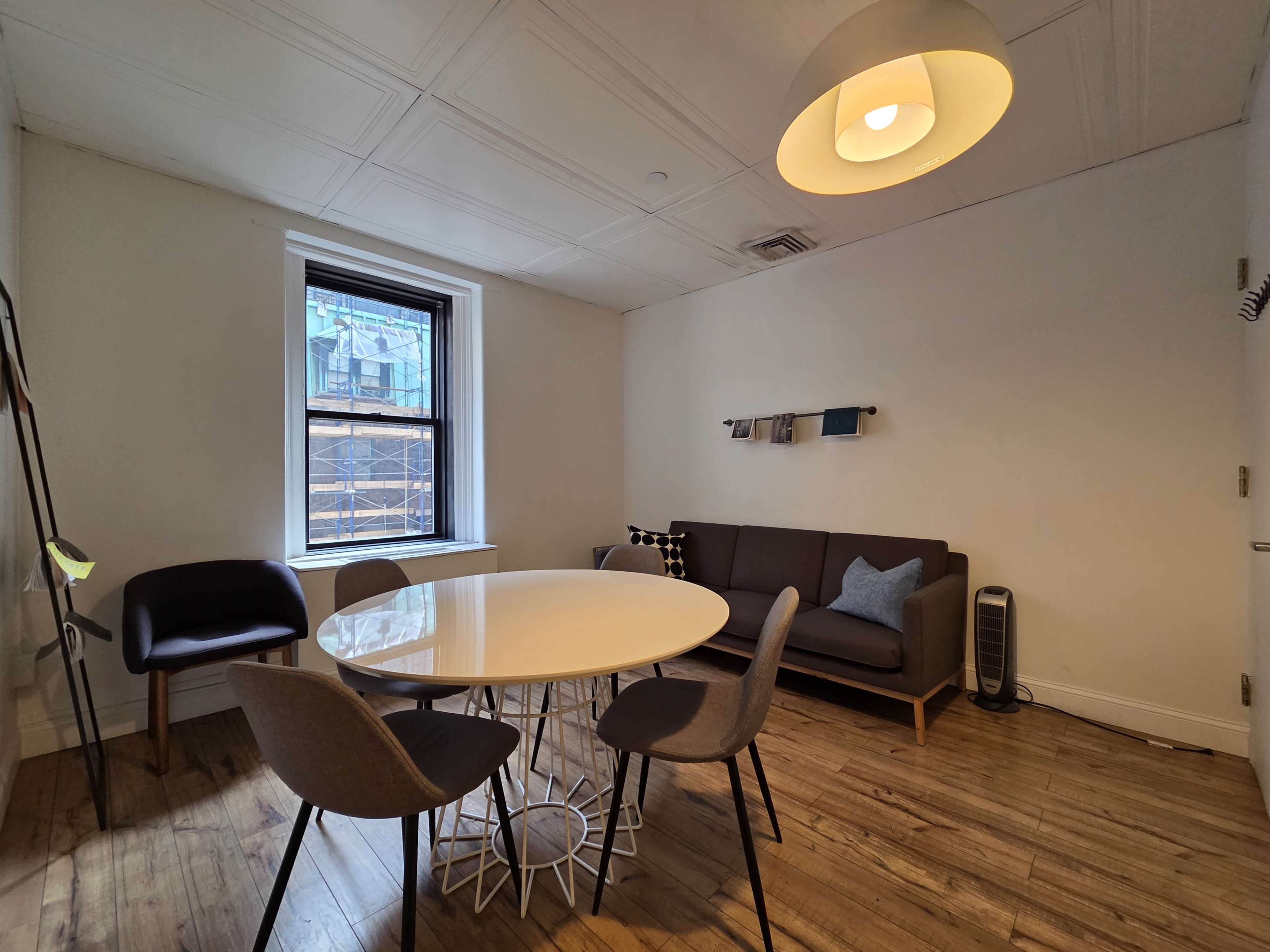 The image shows a simple room featuring a round dining table, four chairs, a sofa against the wall, and a window with curtains.