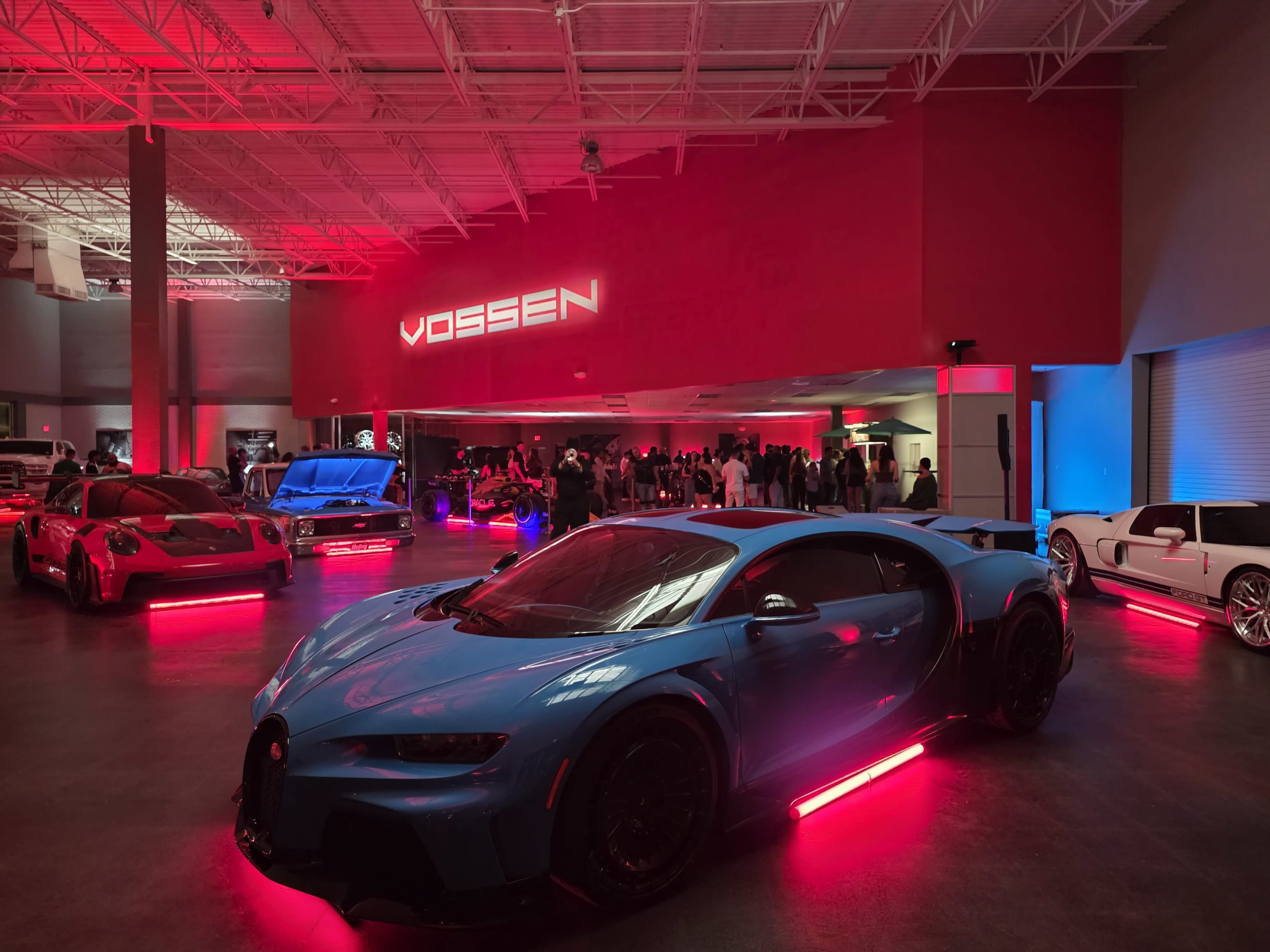The image shows a modern car showroom featuring a blue Bugatti Chiron and several other luxury vehicles, all illuminated by red and blue lighting.