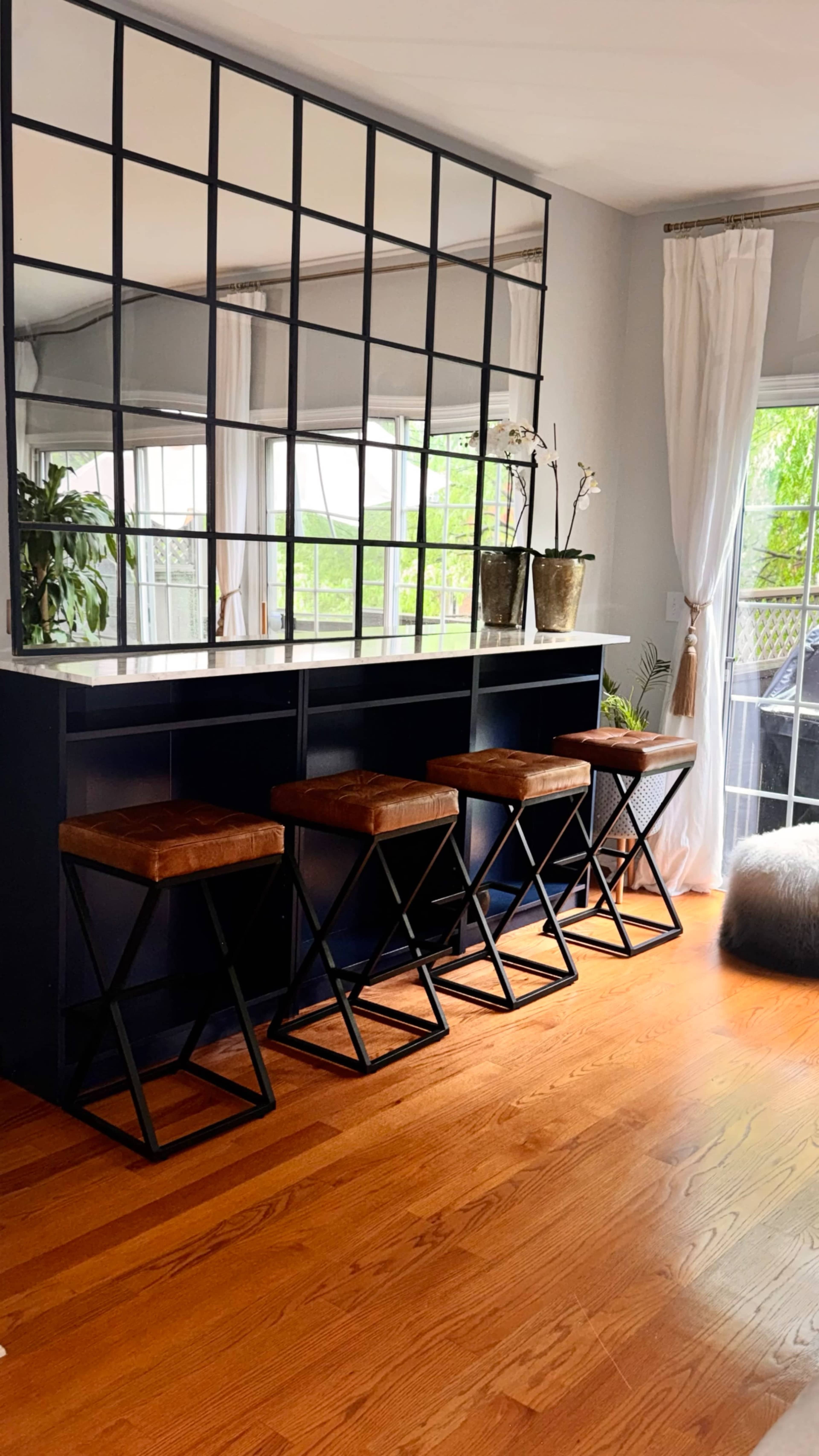 The image shows a modern home interior featuring a bar-style seating area with four stools in front of a large mirror, surrounded by plants and natural light.