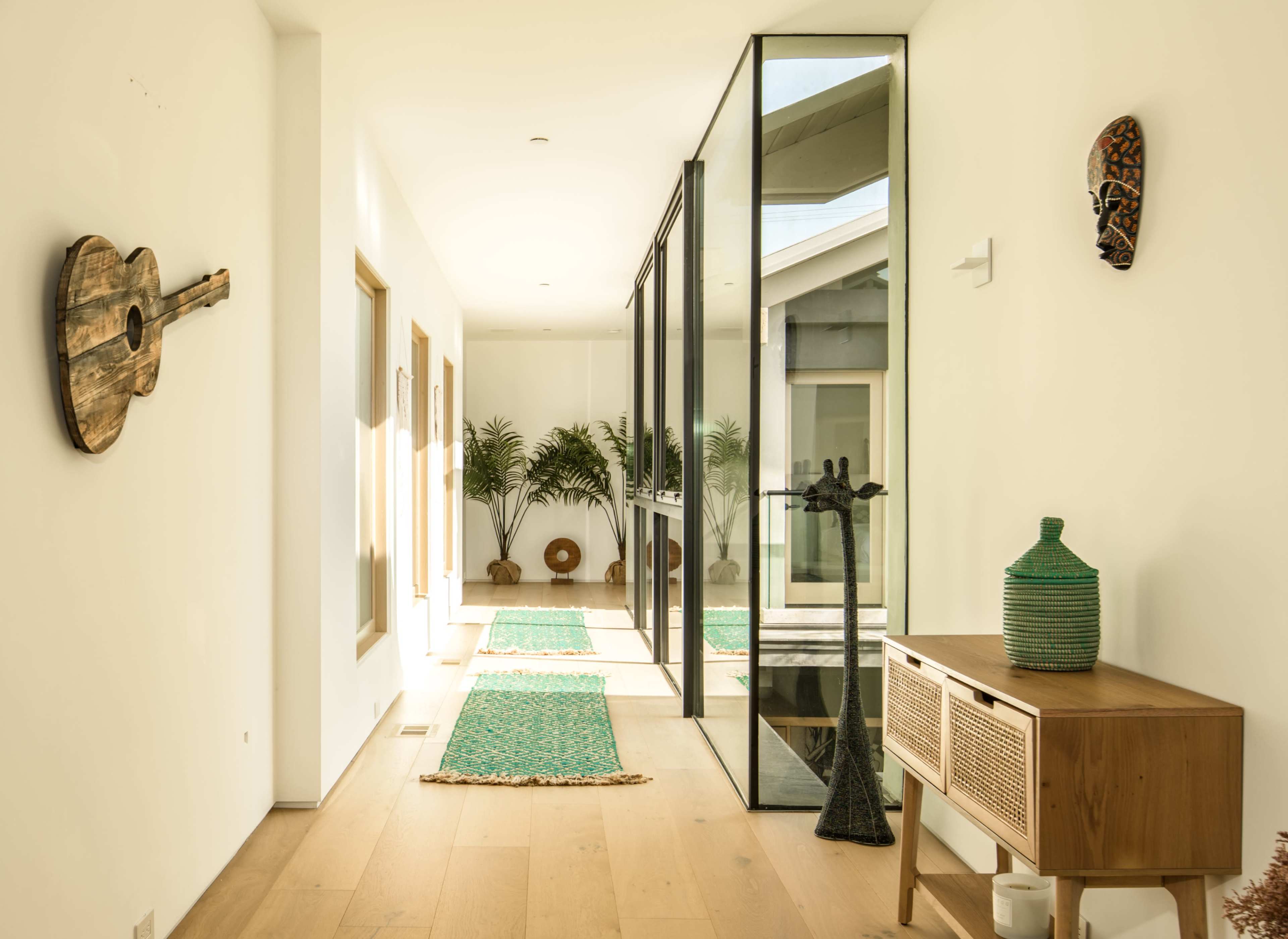 A long, bright hallway features wooden flooring, green rugs, decorative wall art, and large windows leading to a garden.