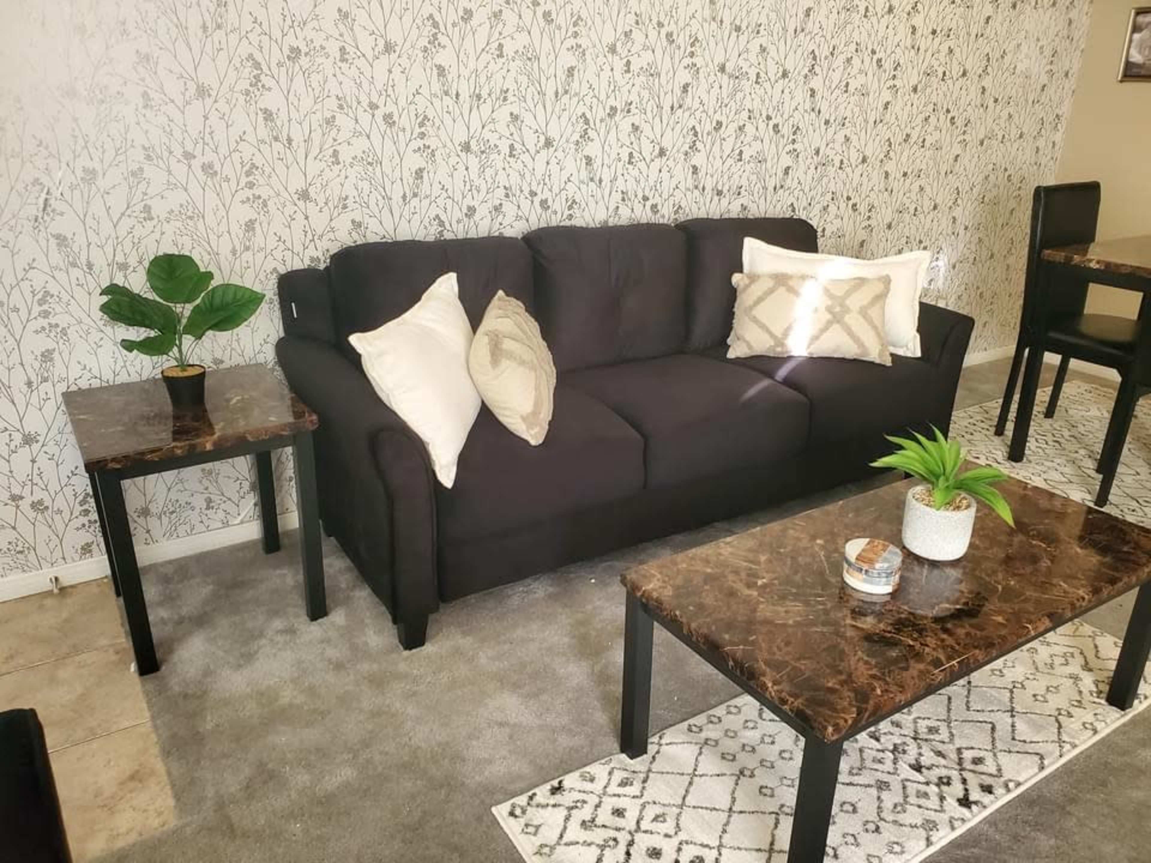 A living room with a black couch, two matching coffee tables with marble tops, and a decorative plant on one table, set against a floral patterned wallpaper.