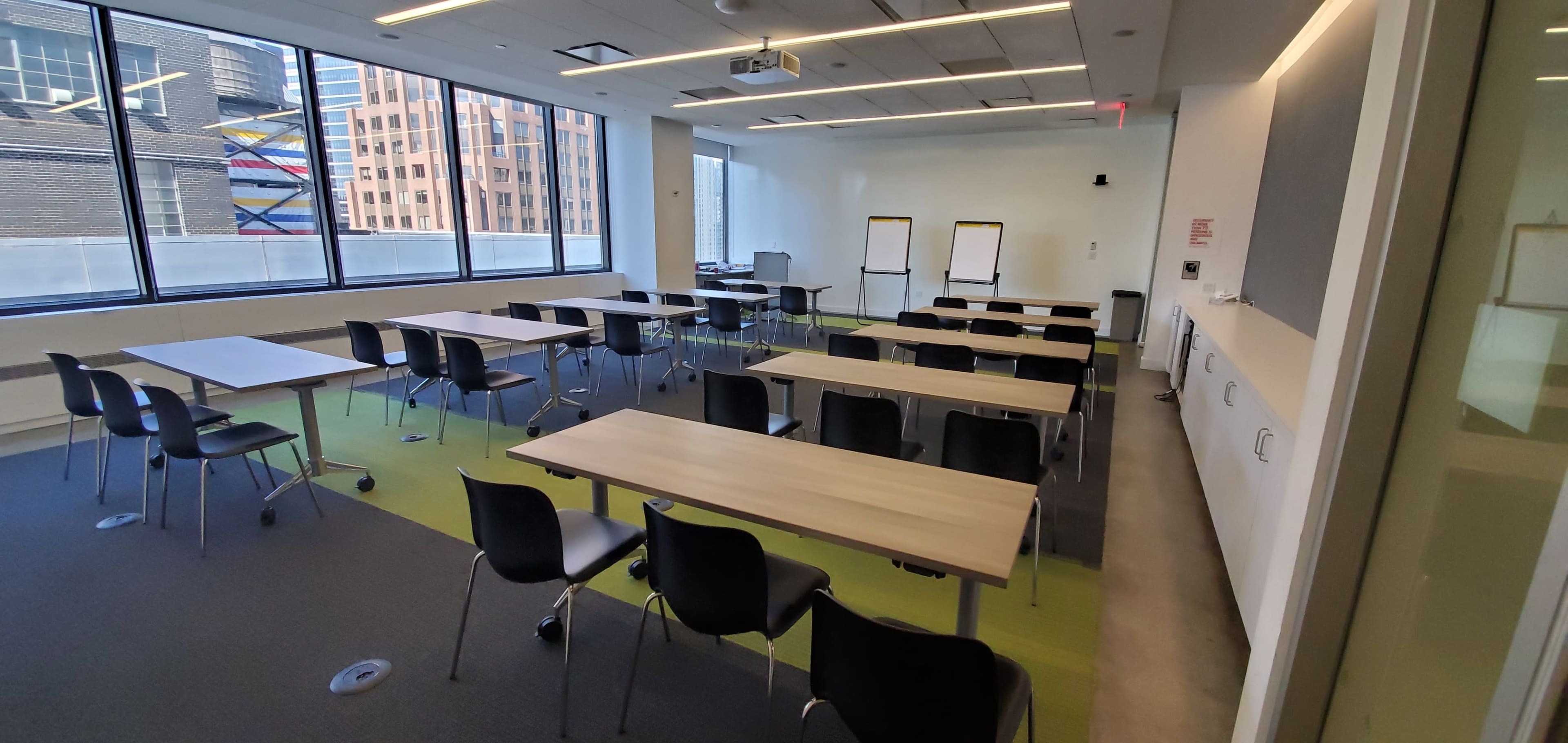 A modern classroom setup features multiple tables and chairs arranged neatly, with large windows overlooking a cityscape.