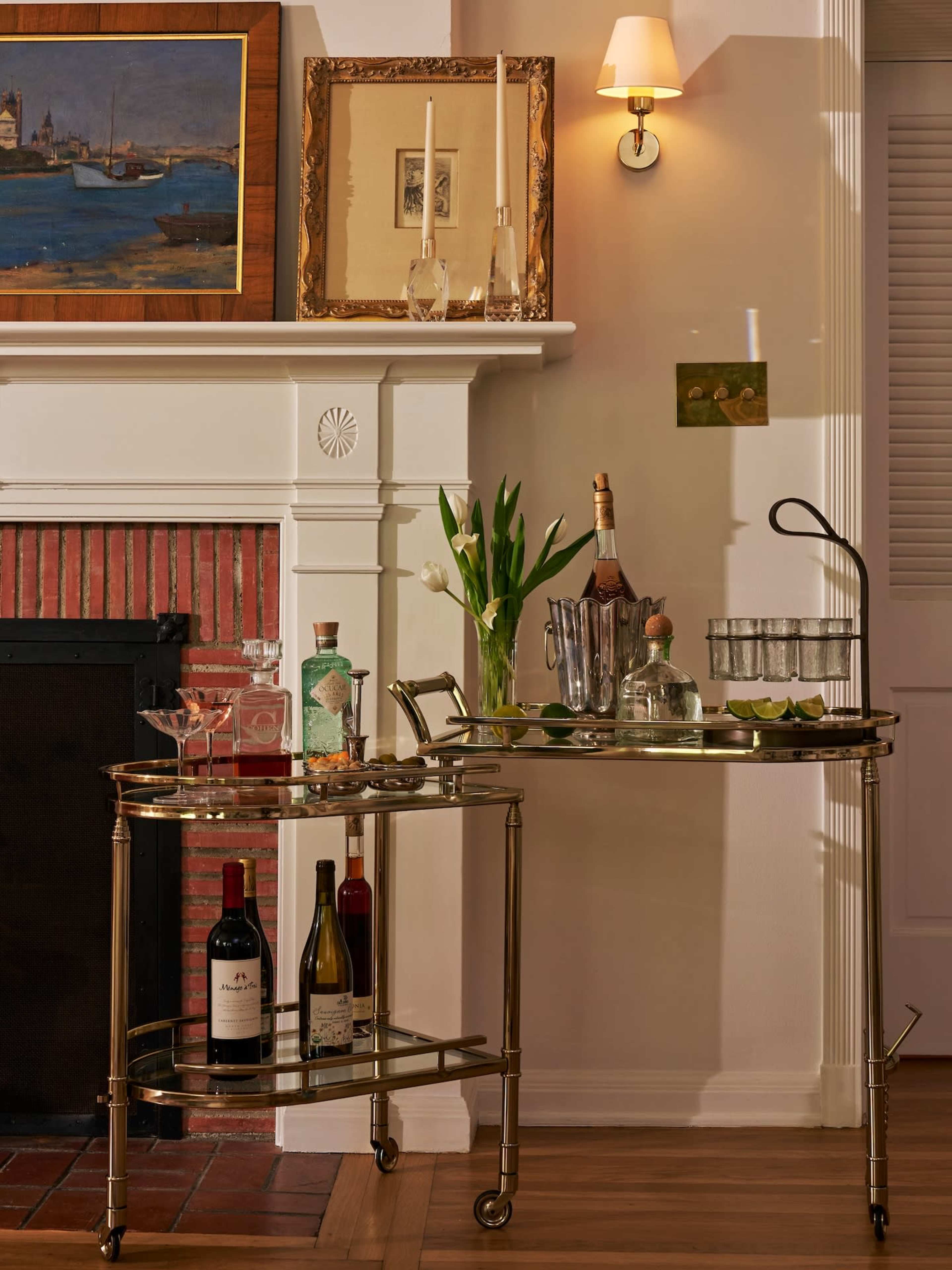 A polished brass bar cart is displayed beside a fireplace, featuring various liquor bottles, glasses, and decorative items, with framed artwork hanging above it.