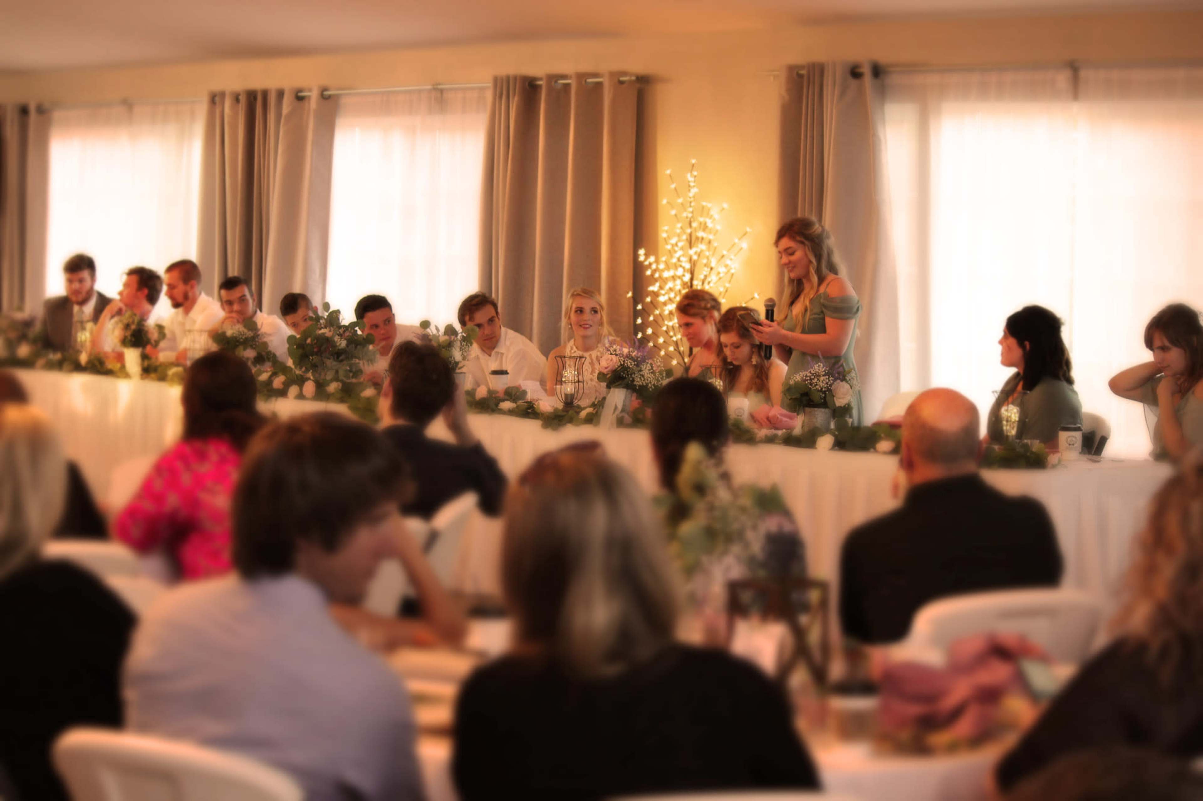 A group of guests is seated at a long table, while one woman stands and speaks, surrounded by decorations and soft lighting.