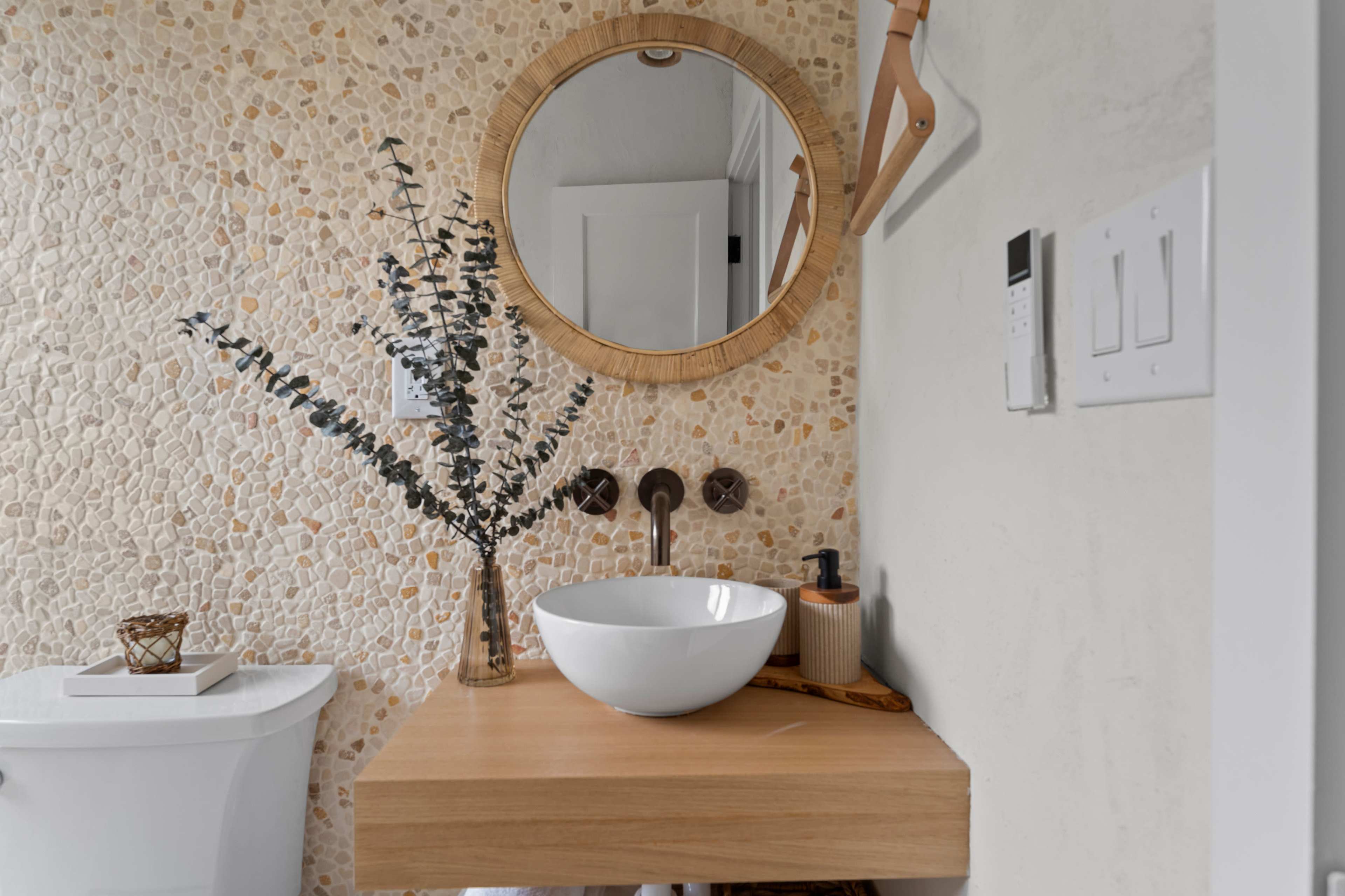 A wooden vanity with a round mirror and decorative vase is positioned against a textured stone wall in a bathroom.