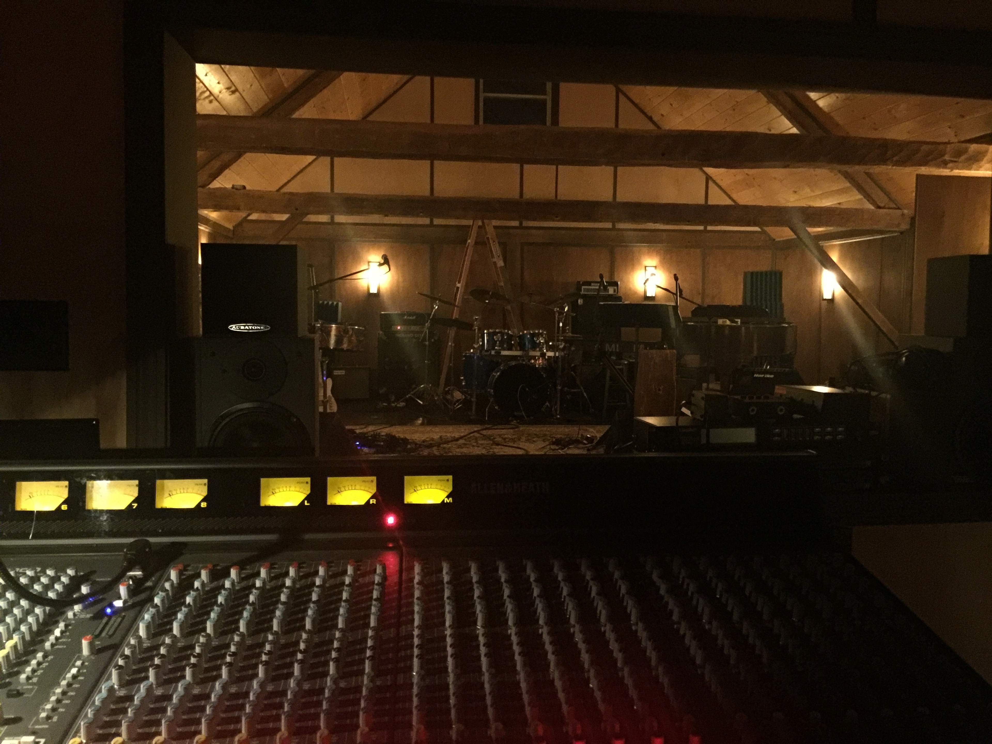 A recording studio features a sound mixing console in the foreground, with a well-equipped drum set and various instruments visible in the background.