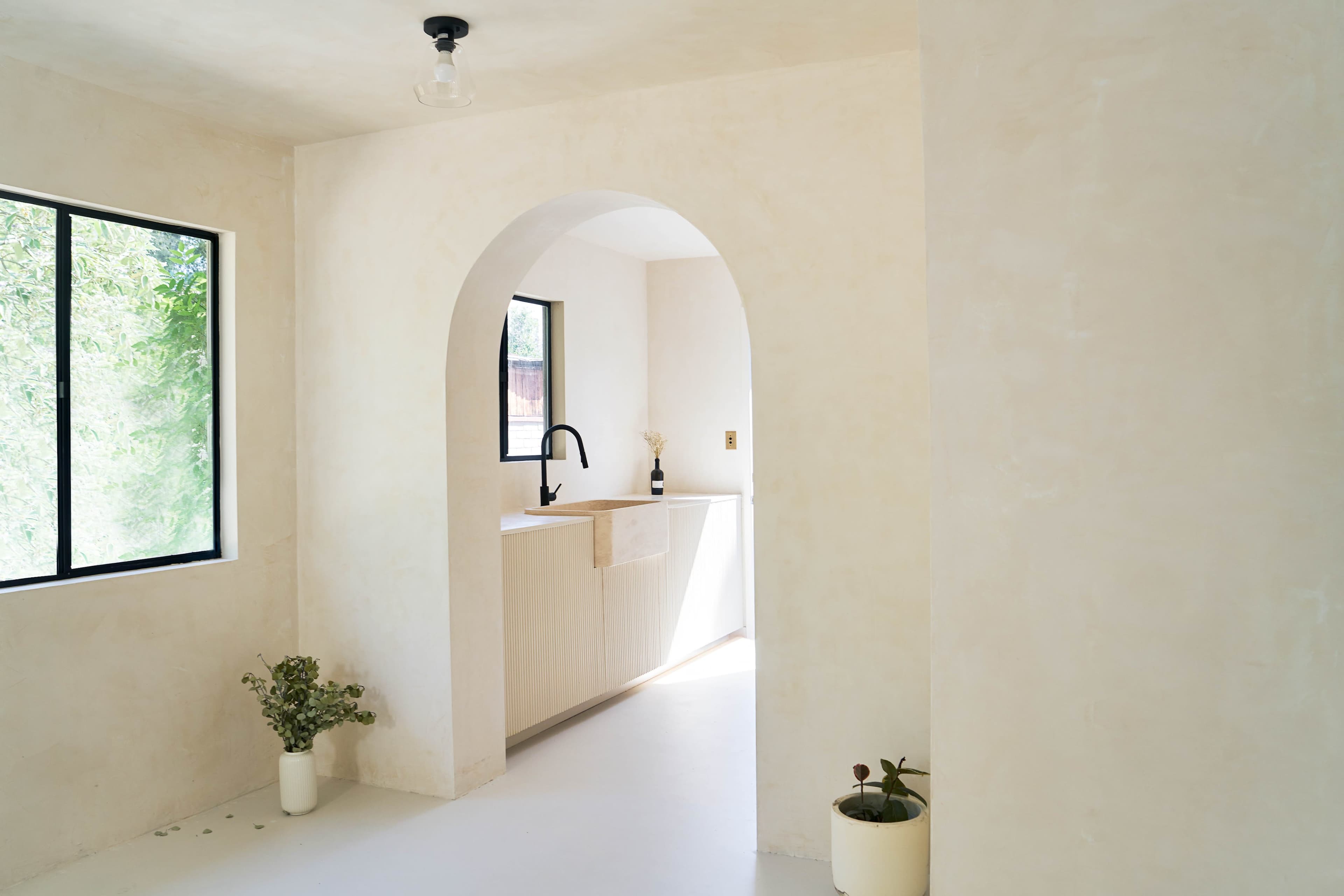 A minimalist interior with a curved archway leading to a kitchen area, featuring large windows and light-colored walls.