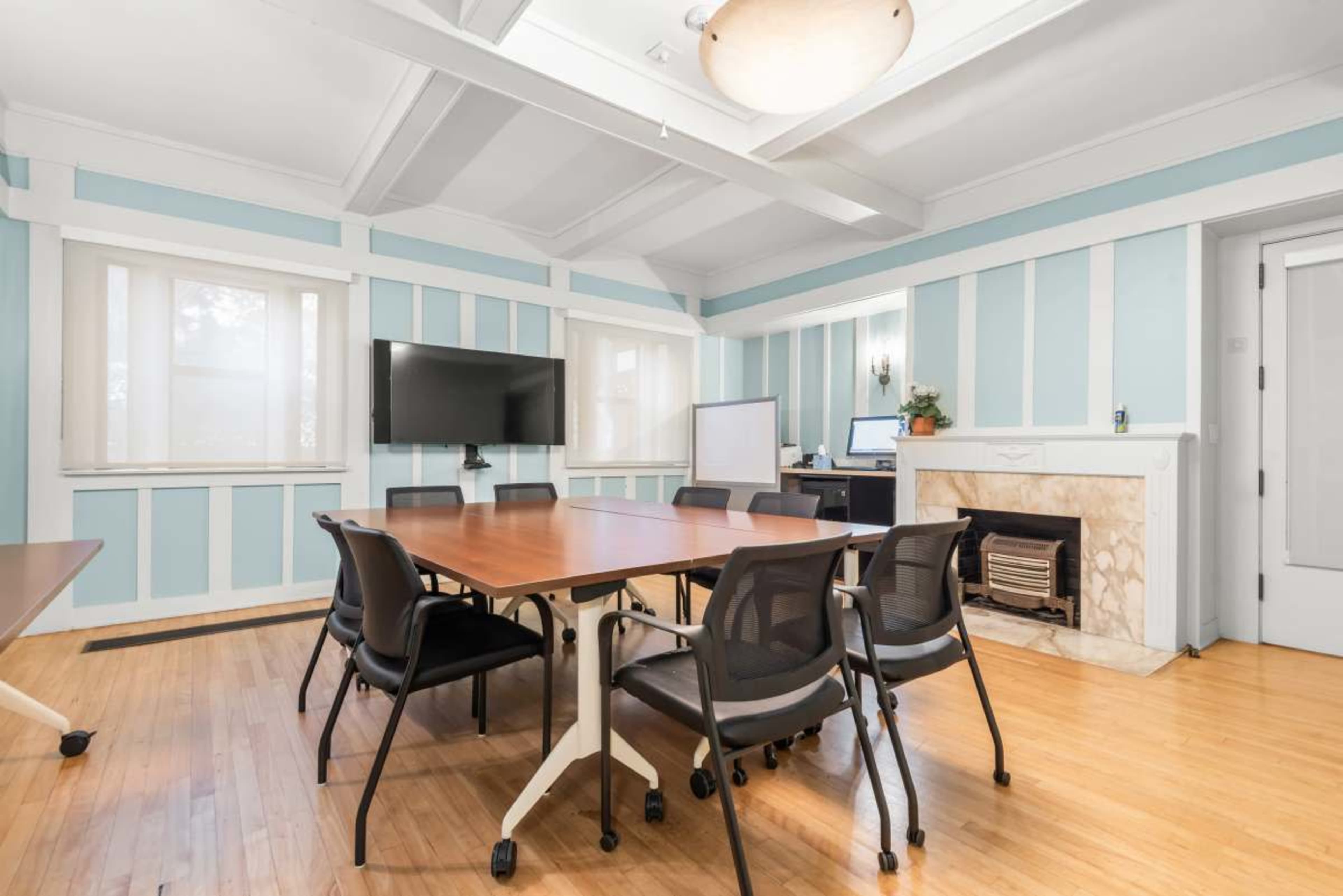 The image shows a well-lit conference room featuring a large wooden table surrounded by chairs, a wall-mounted TV, and a fireplace.