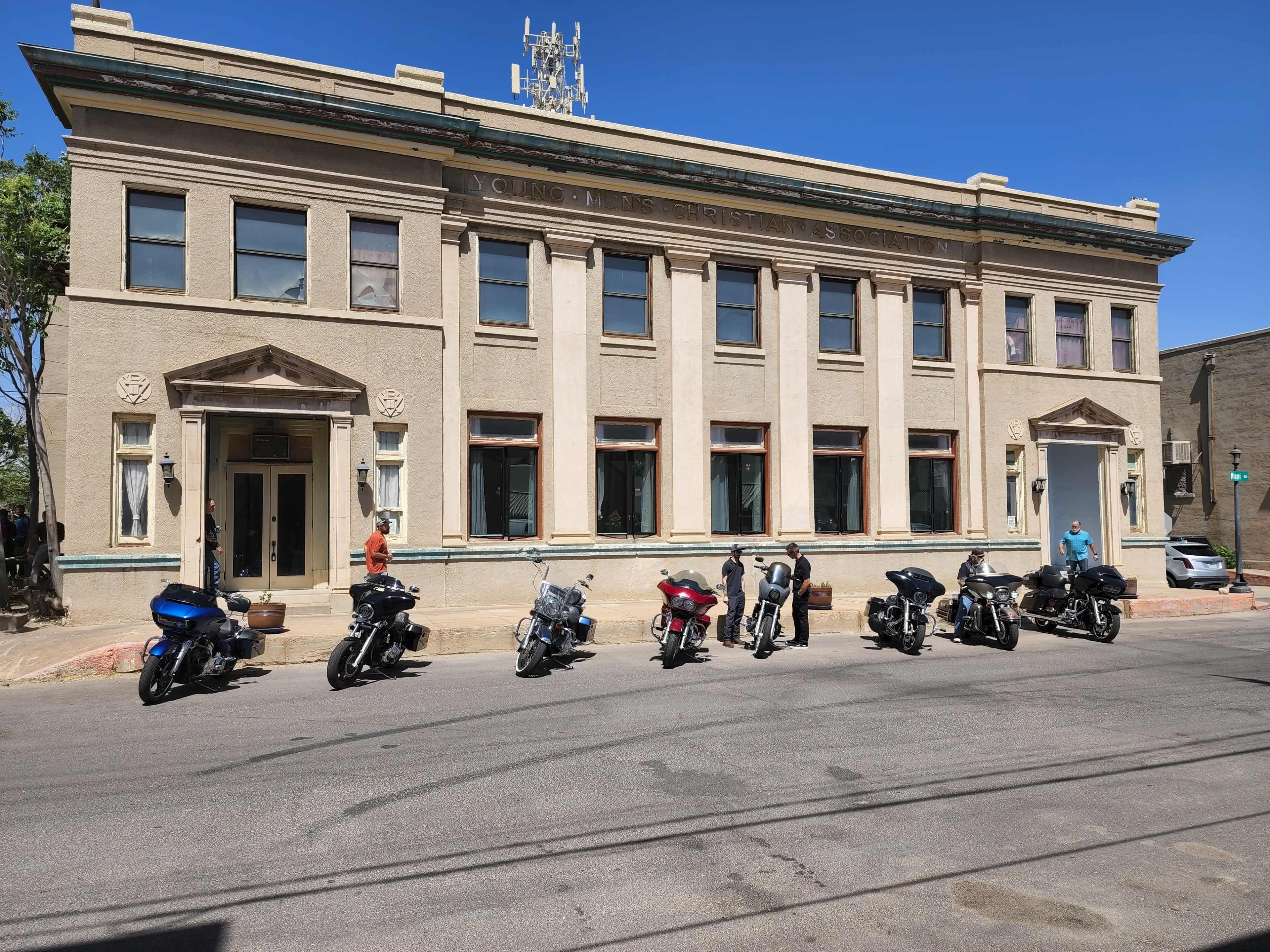 A row of parked motorcycles is lined up in front of a historic, two-story building with a telecommunications tower on its roof.
