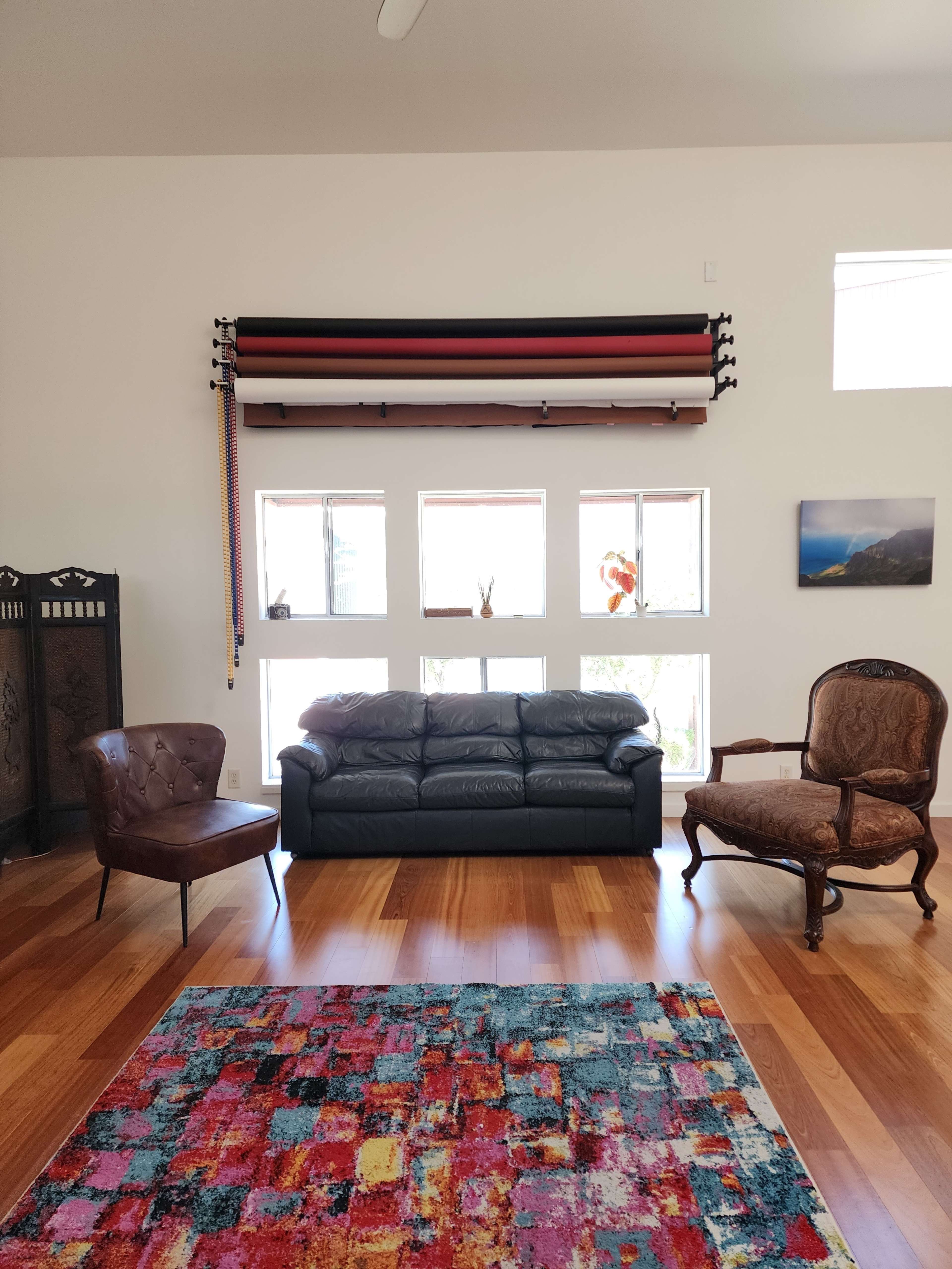 A living room features a black leather sofa, a vintage brown chair, and a patterned rug, with large windows that allow natural light to enter.