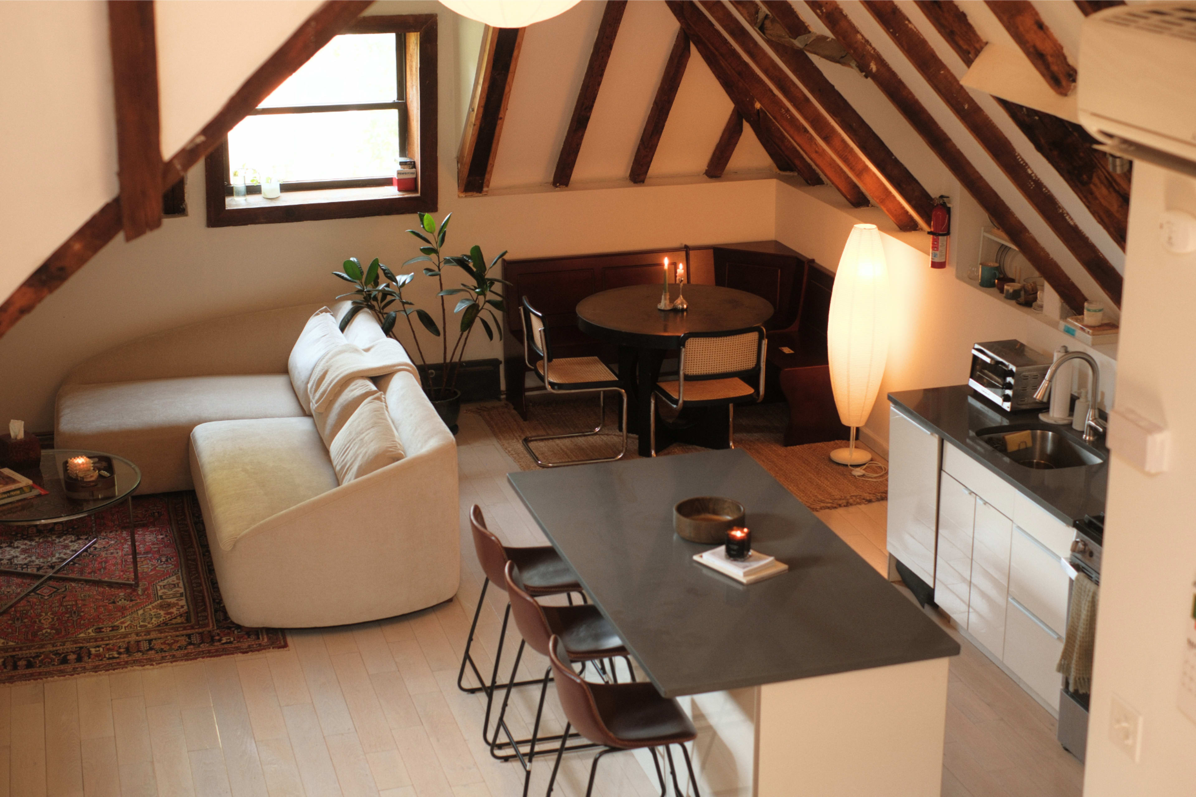 The image shows a cozy attic apartment featuring a living area with a sofa, a circular dining table, and a modern kitchen with bar stools.