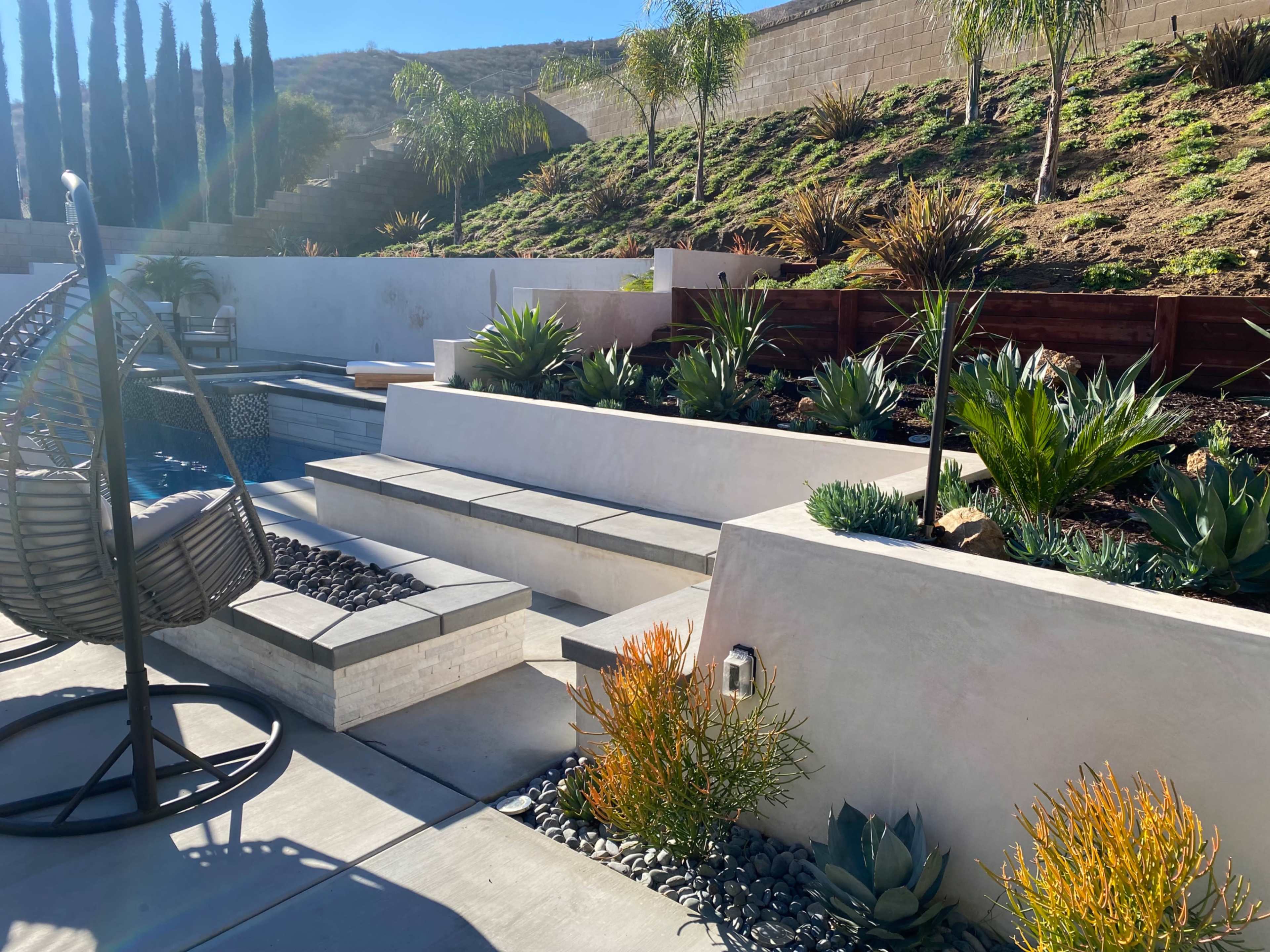 A modern outdoor area with a swimming pool, tiered stone steps, and various plants including agaves and grasses.