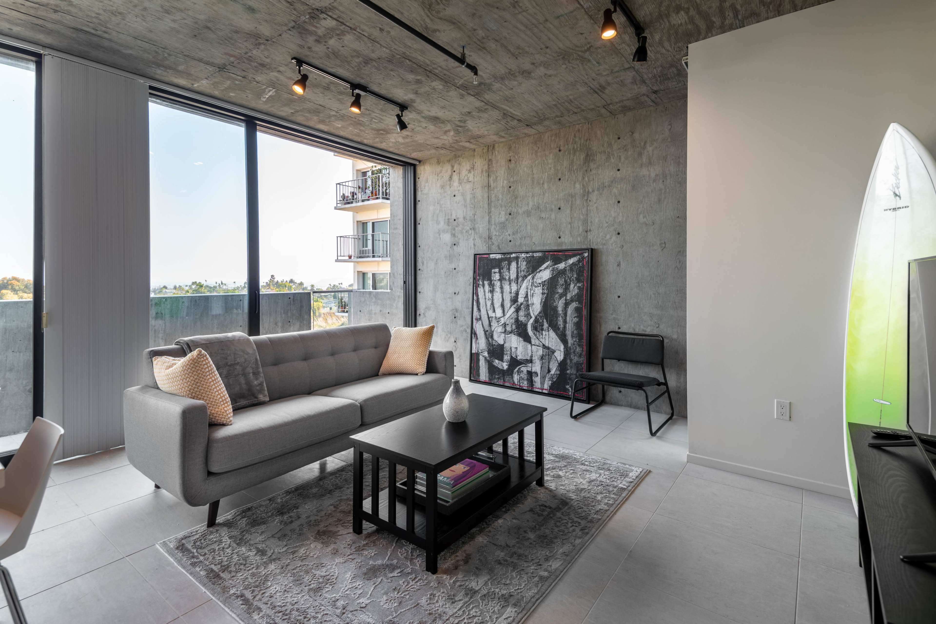 A modern living room features a gray sofa, a black coffee table, and a large window with a view of a balcony and surrounding area.