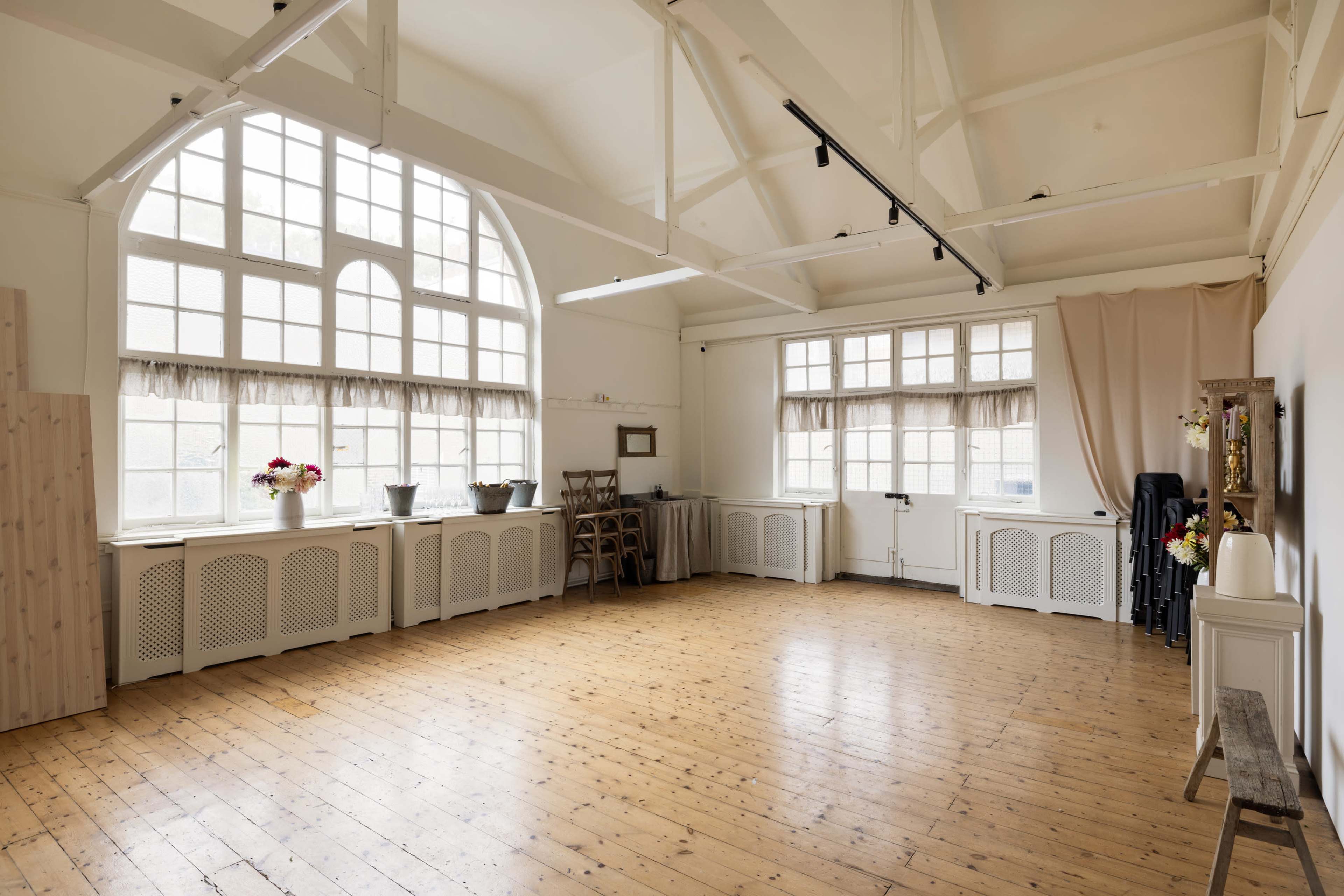 The image shows an empty room with wooden floors, large arched windows allowing natural light, and minimal furnishings including folding chairs and decorative elements.