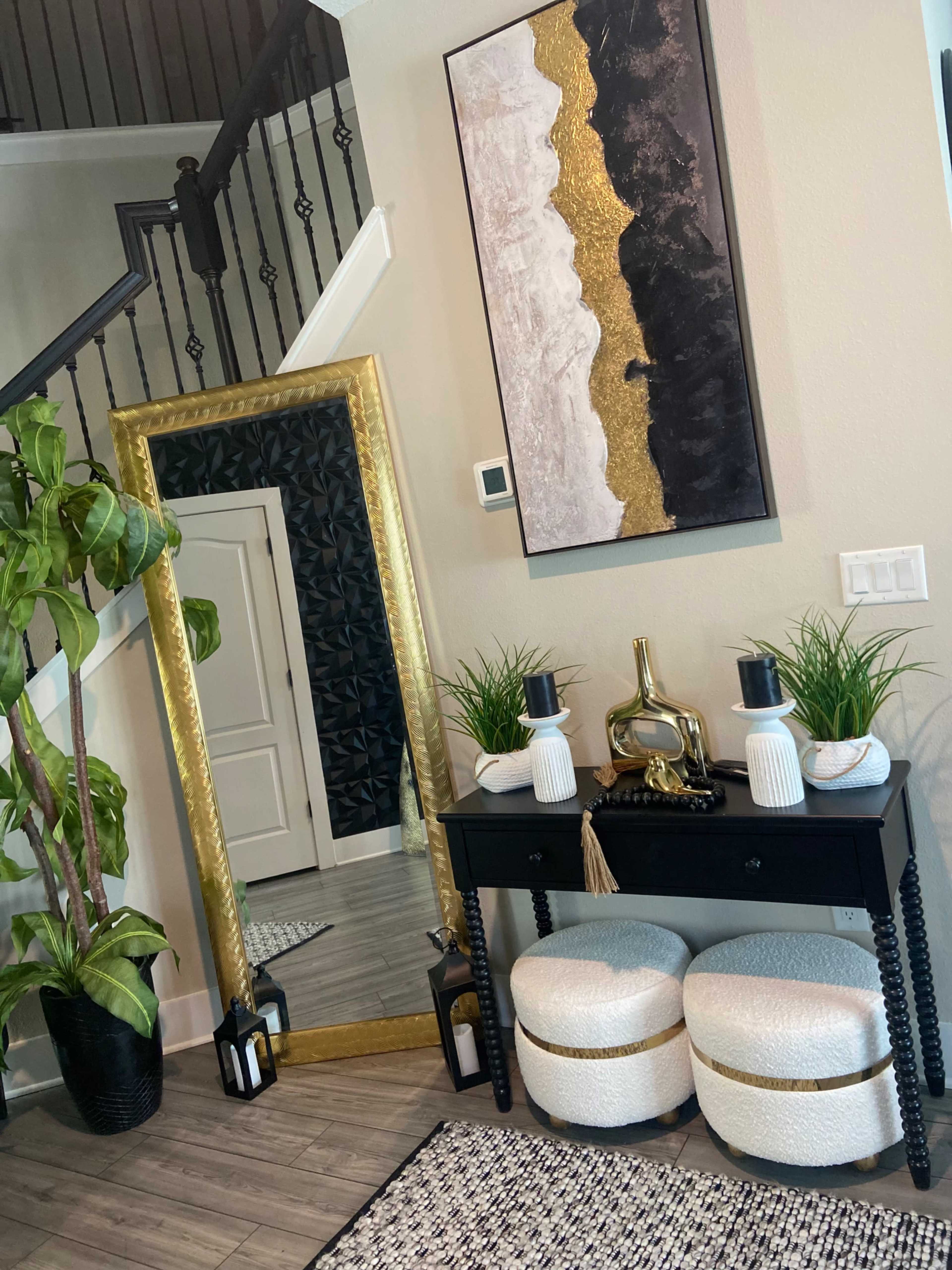 The image shows a stylish entryway featuring a large gold-framed mirror, a black console table with decorative plants and candles, and two round poufs.