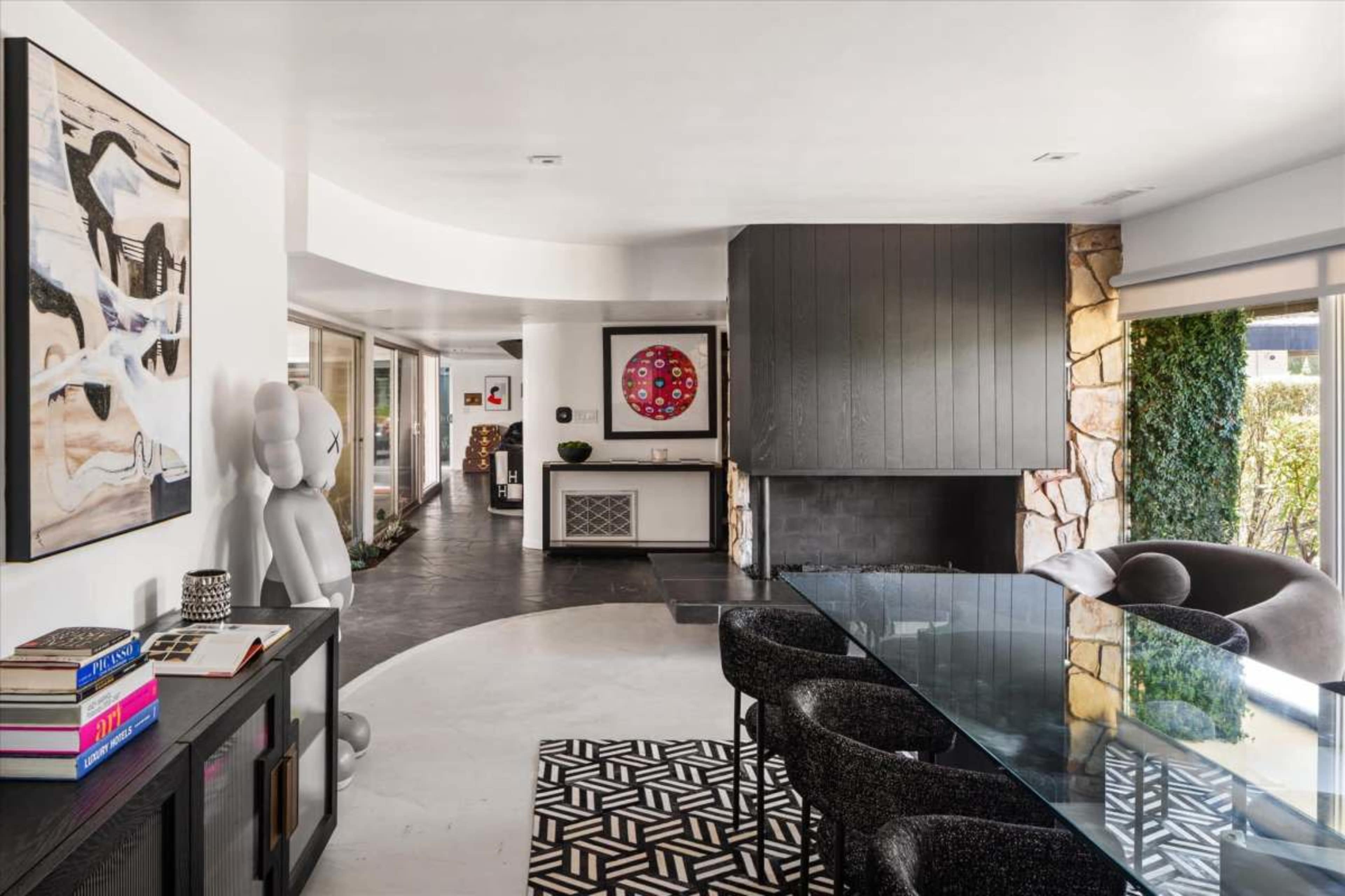 The image shows a modern interior space featuring a glass dining table surrounded by black chairs, with a fireplace and artwork on the walls.