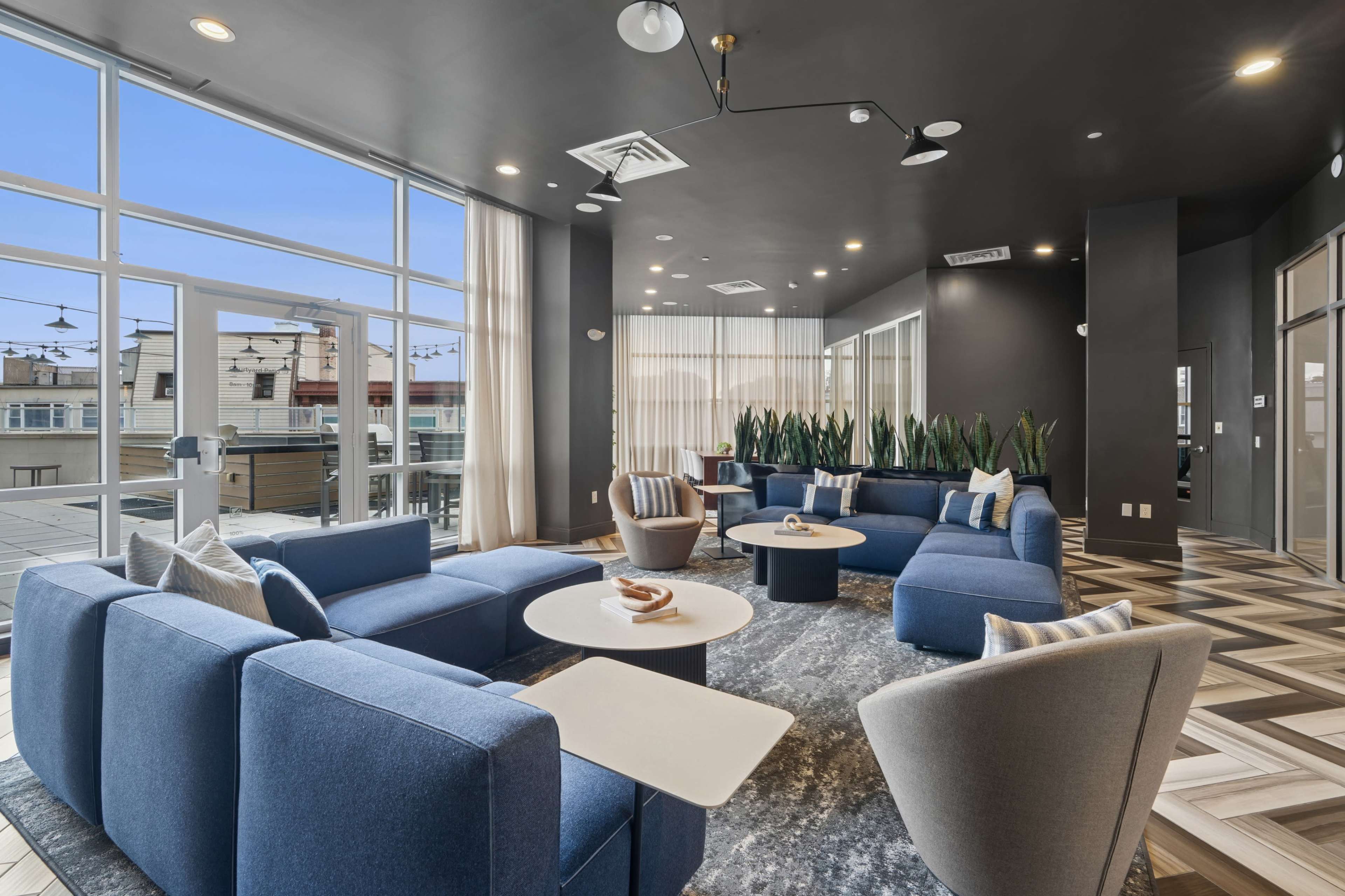 The image shows a modern lounge area with distinct blue furniture, large windows, and plants, designed for relaxation and socializing.