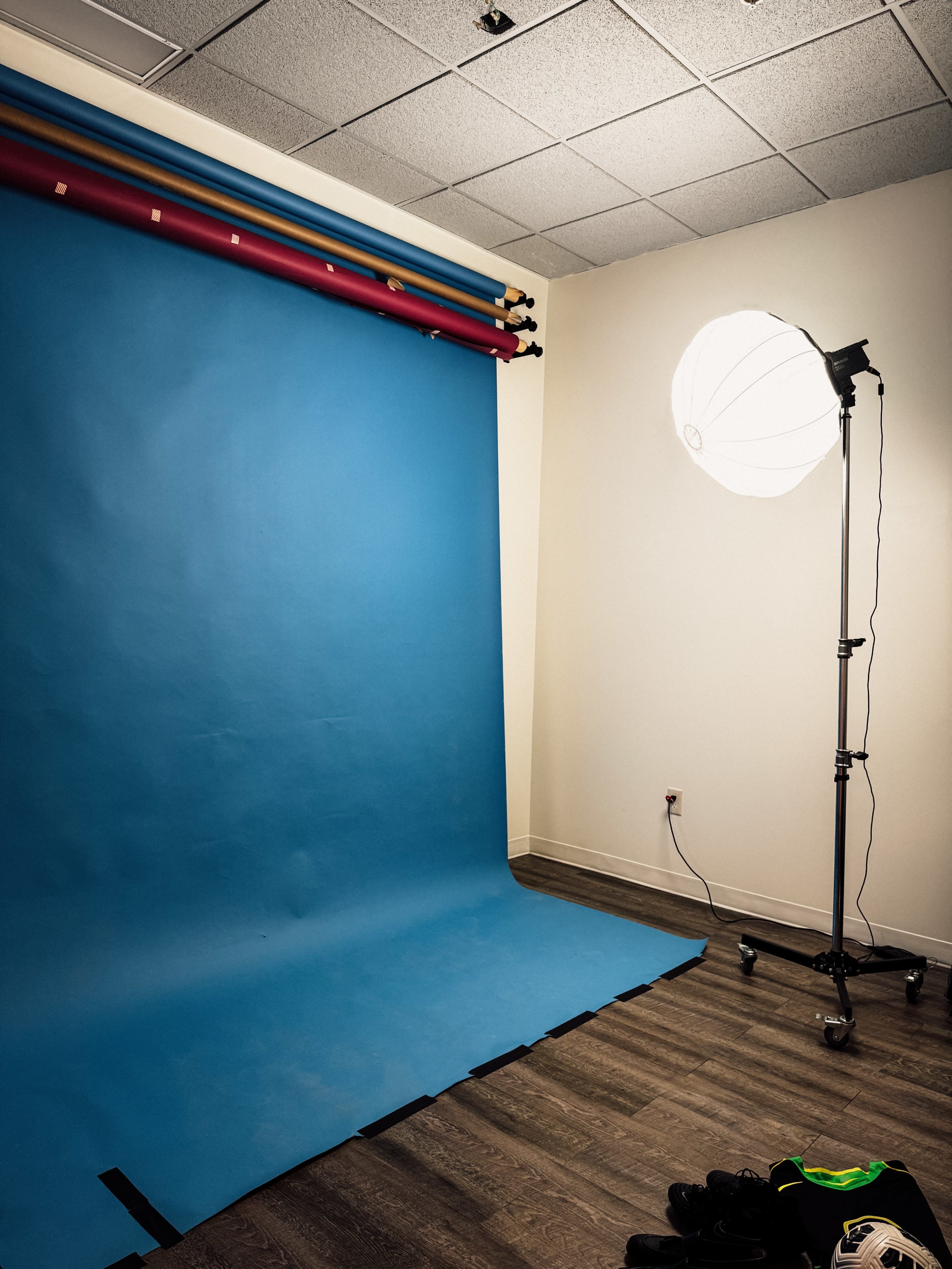 The image shows a small studio setup with a blue backdrop, a light source on a stand, and some clothing items on the floor.