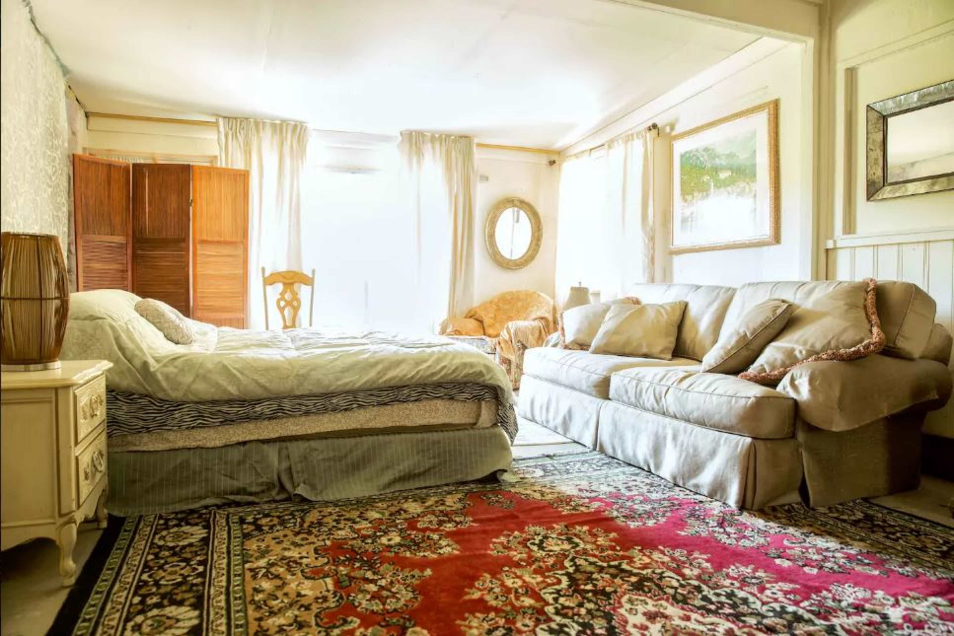 A cozy bedroom features a bed, a couch, and large windows allowing natural light to fill the space, with a decorative rug on the floor.
