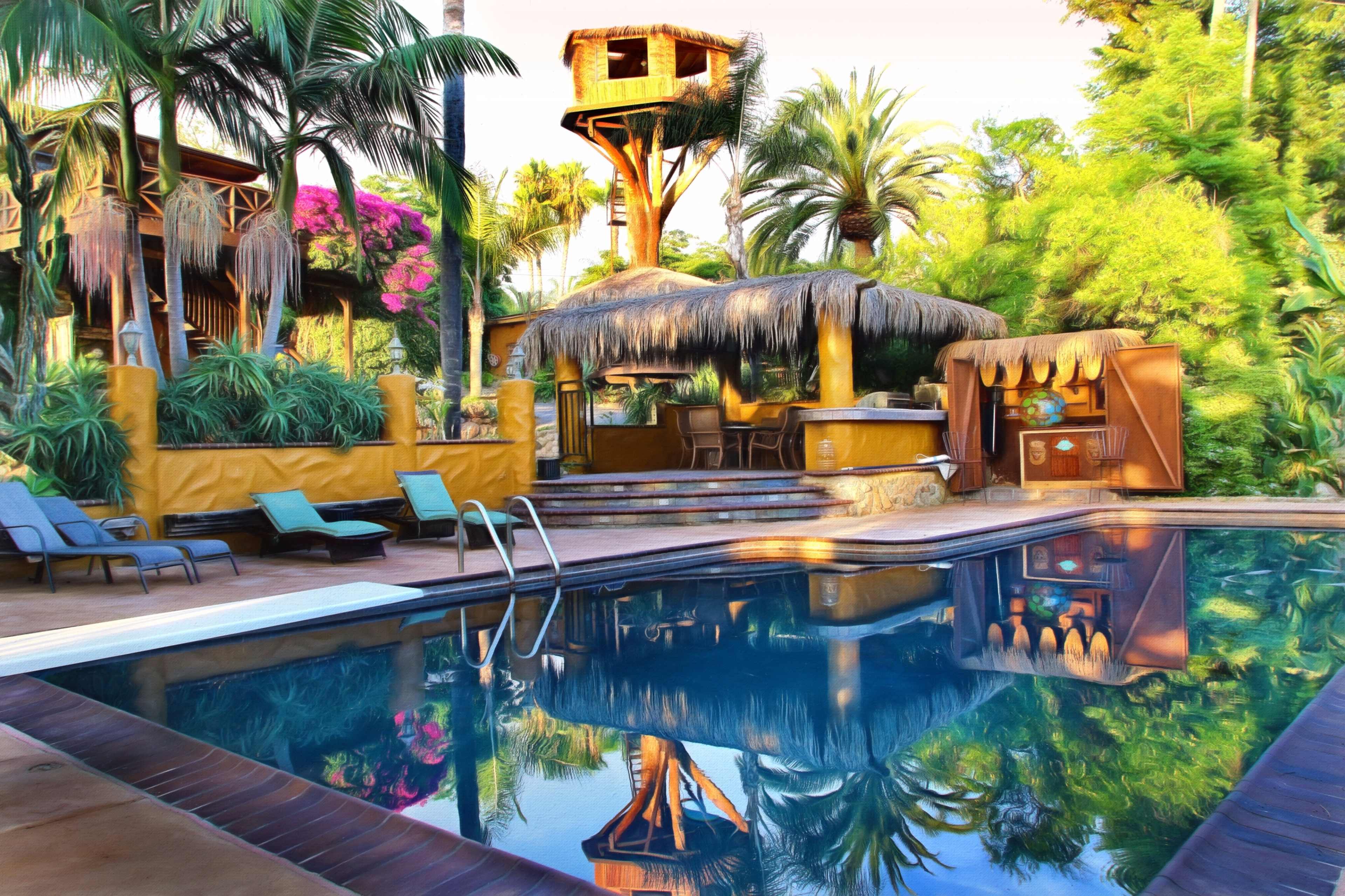A swimming pool reflects a tropical landscape featuring a thatched roof bar and a raised treehouse surrounded by palm trees and vibrant plants.