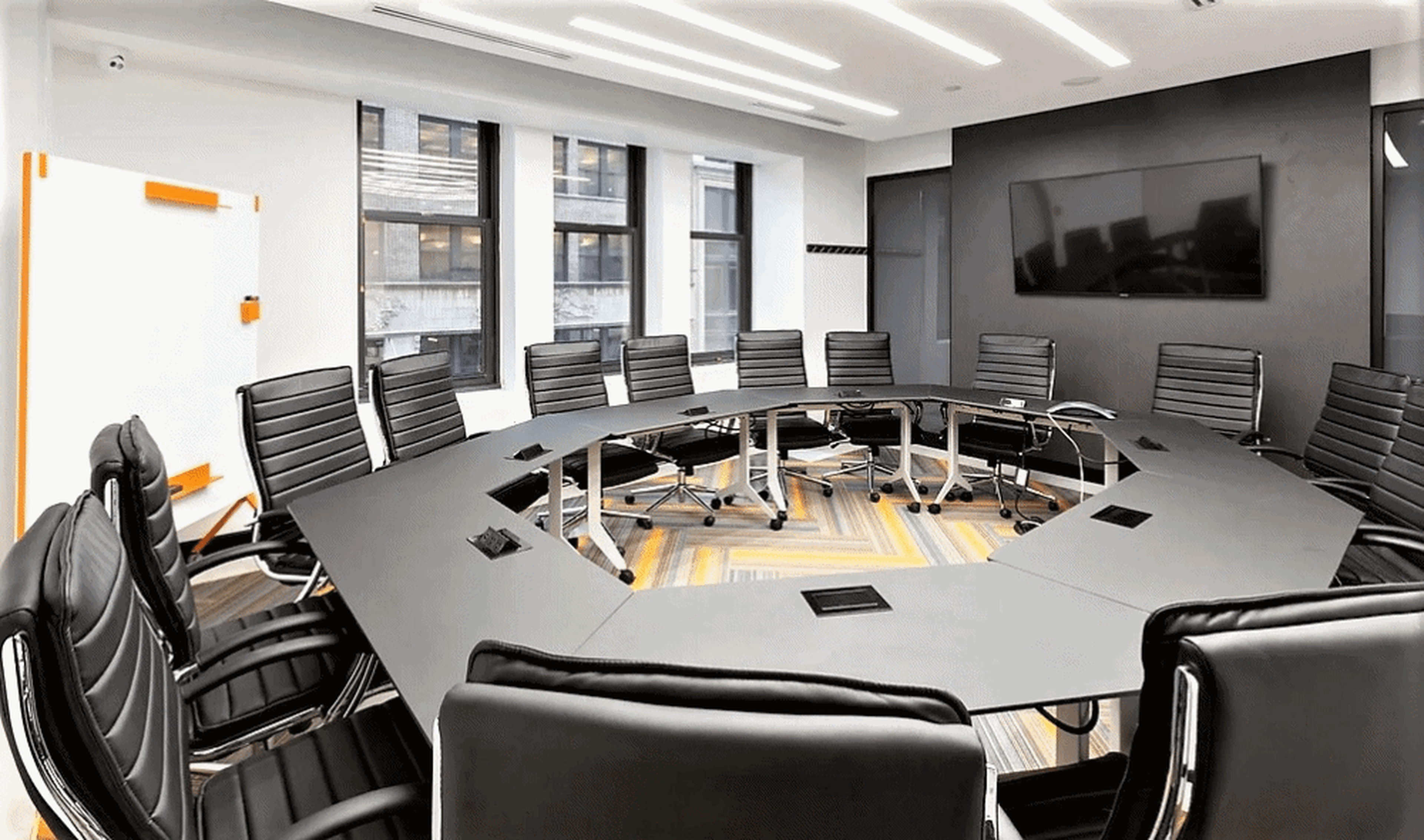 A modern conference room features a large, oval table surrounded by black executive chairs, with a wall-mounted TV and large windows providing natural light.
