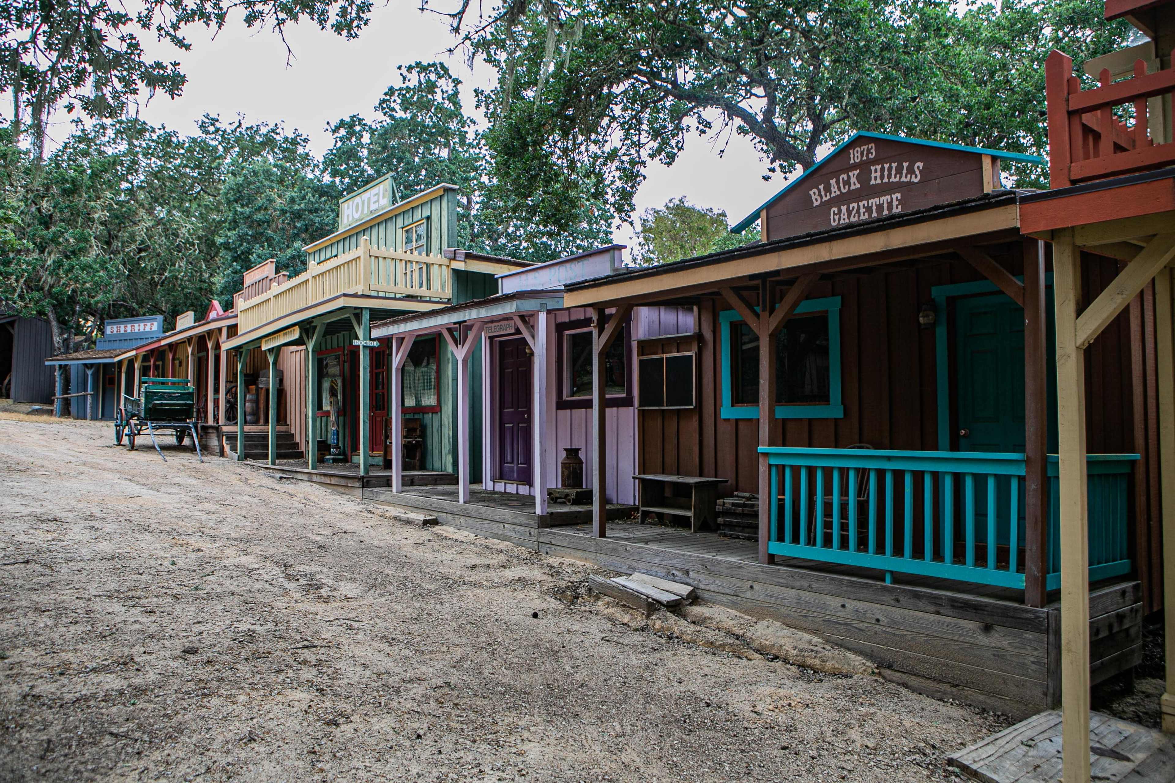 A row of historic wooden buildings with colorful facades lines a dirt street in a rural setting.