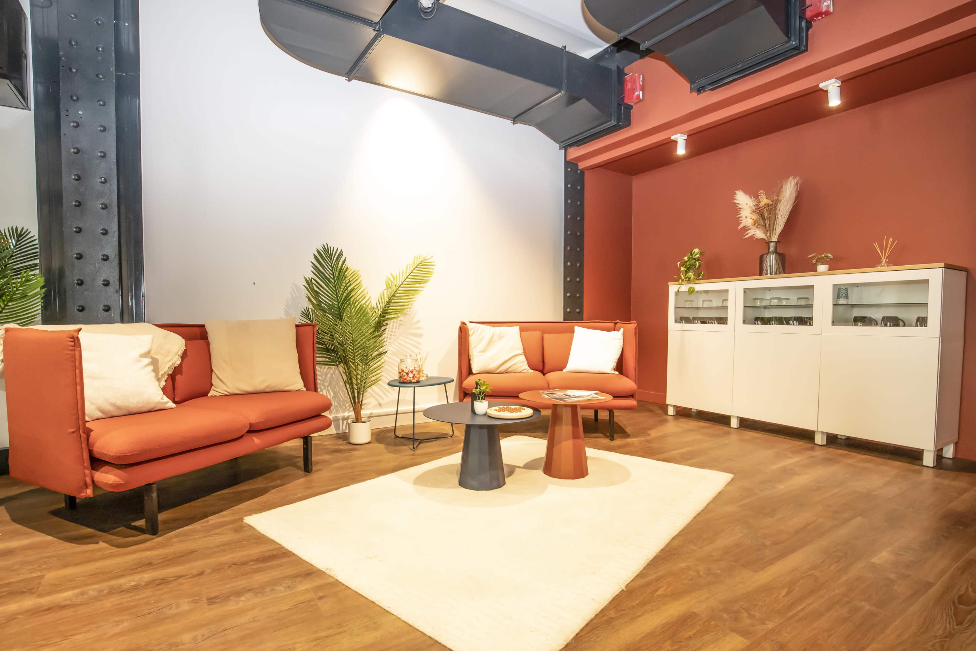 The image shows a modern lounge area with two red couches, a round black and white coffee table, and a small plant beside a white cabinet against a wall.