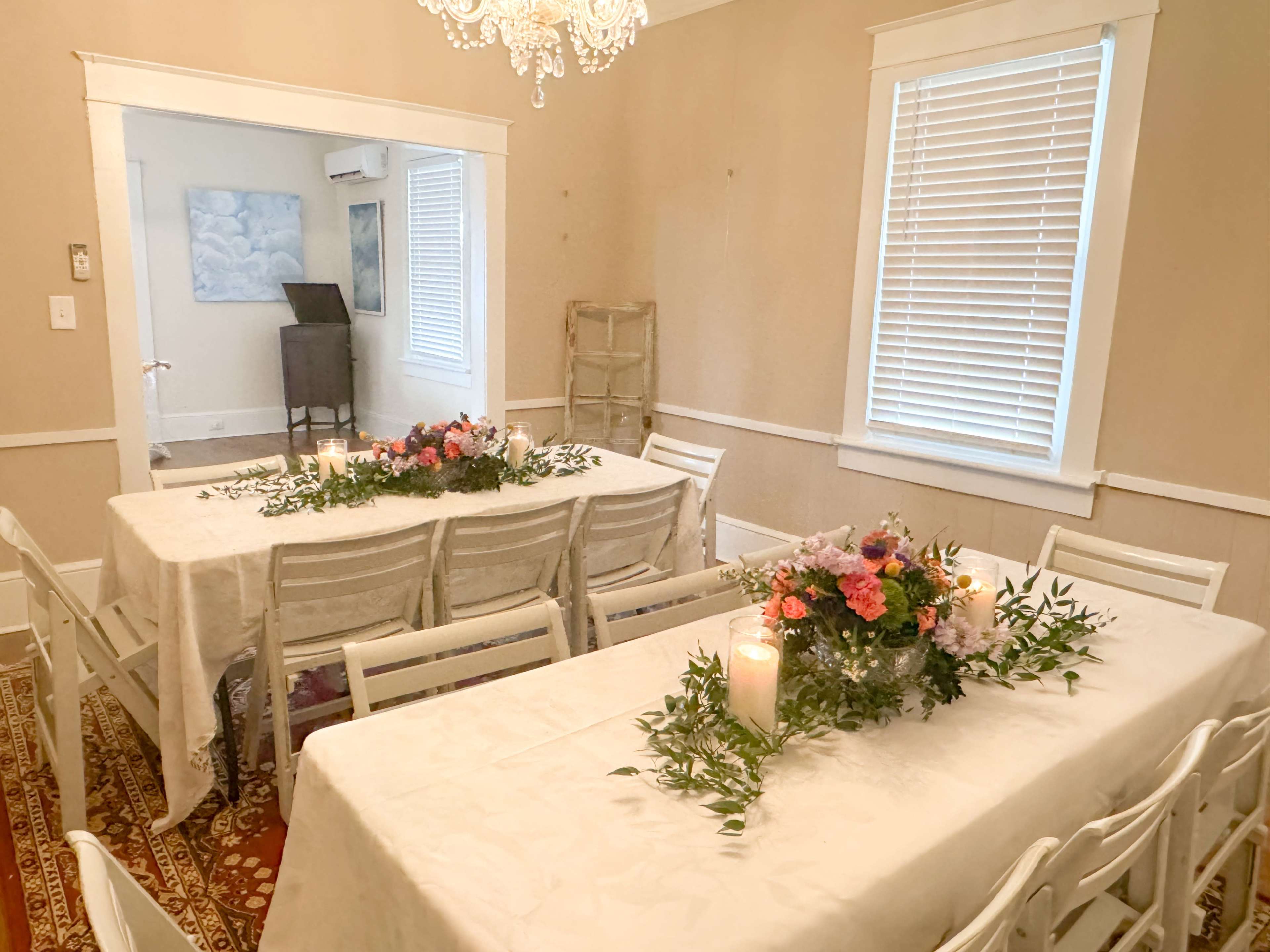 A dining area features two long tables adorned with floral centerpieces and candles, surrounded by white chairs.