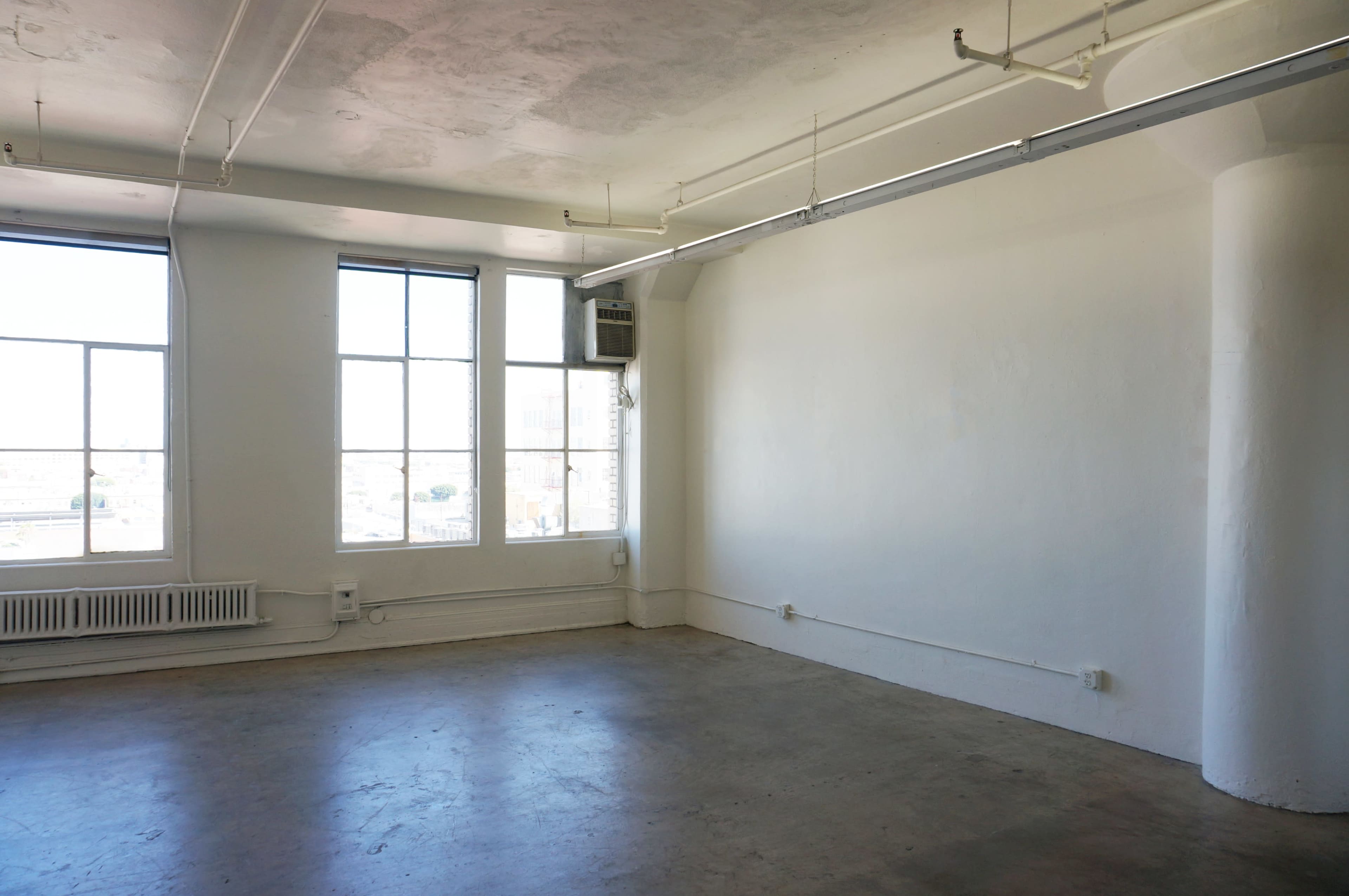 The image shows a spacious, empty room with concrete flooring, large windows, and white walls.