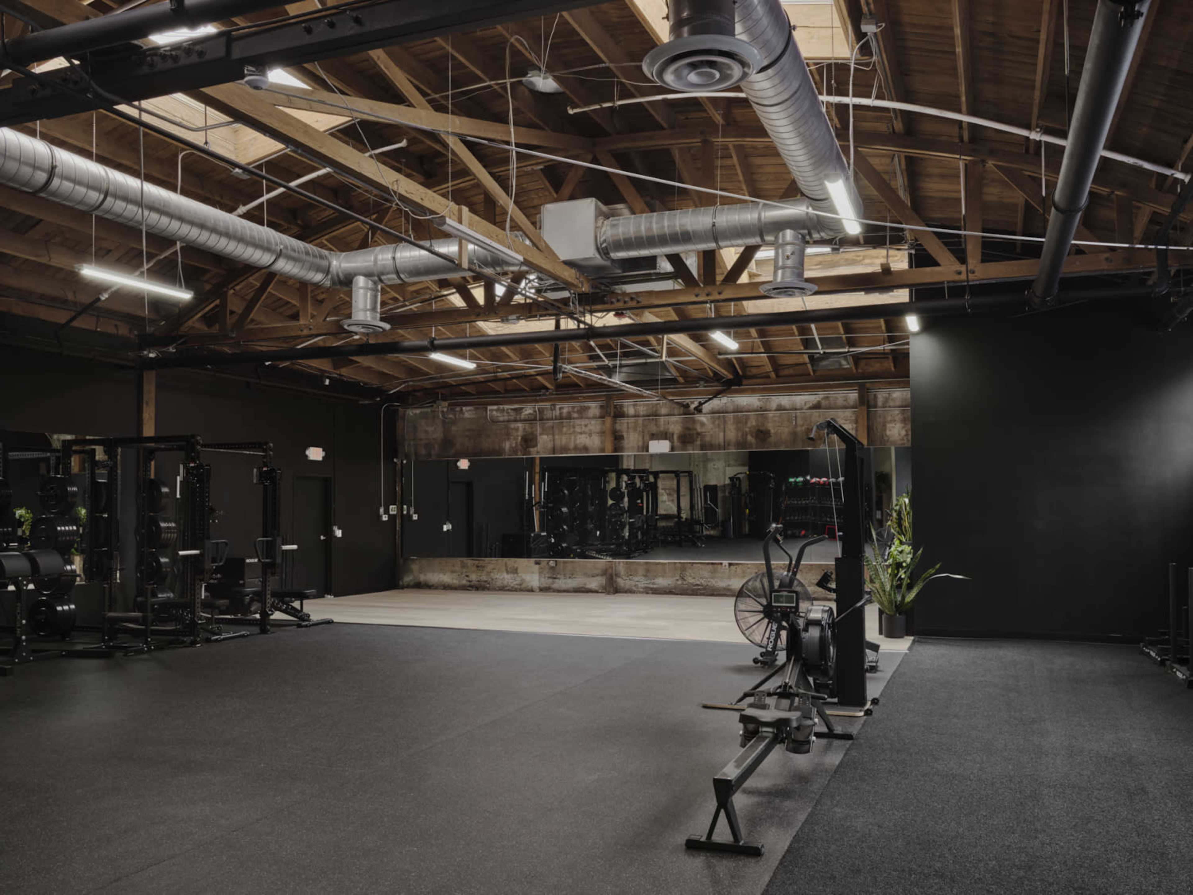 A spacious gym with various workout equipment, including weight racks and rowing machines, set against a backdrop of exposed wooden beams and industrial-style lighting.