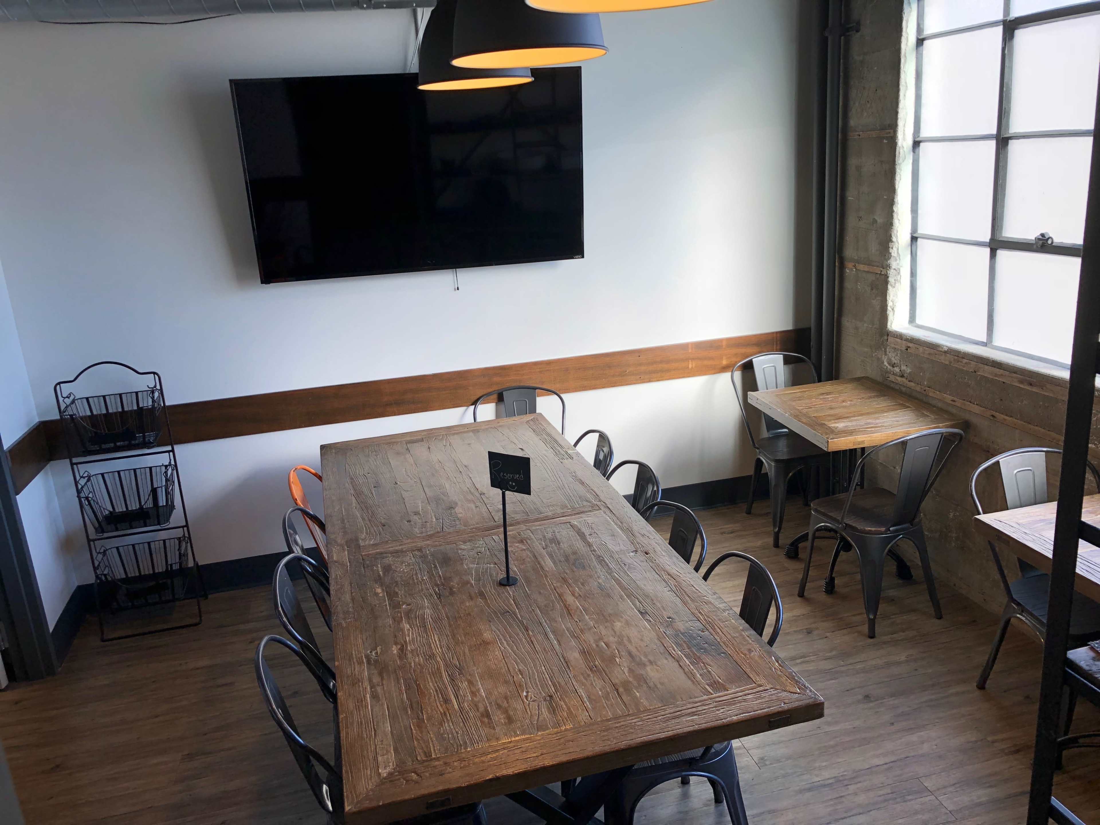 A spacious room with wooden tables, metal chairs, a large wall-mounted TV, and large windows allowing natural light.