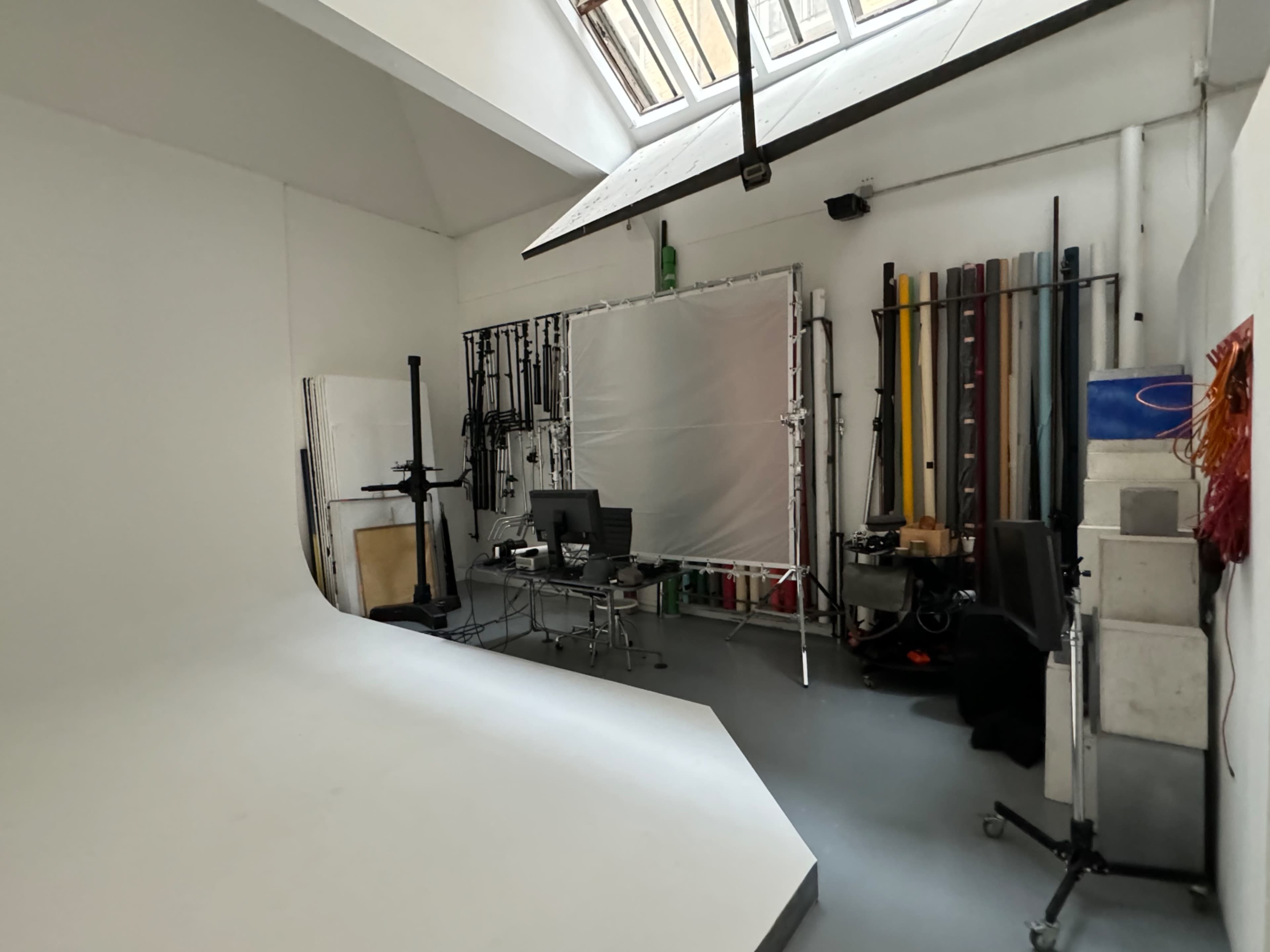 The image shows a photography studio featuring a white curved backdrop, a desk with equipment, and various colorful backdrops stored vertically along the wall.