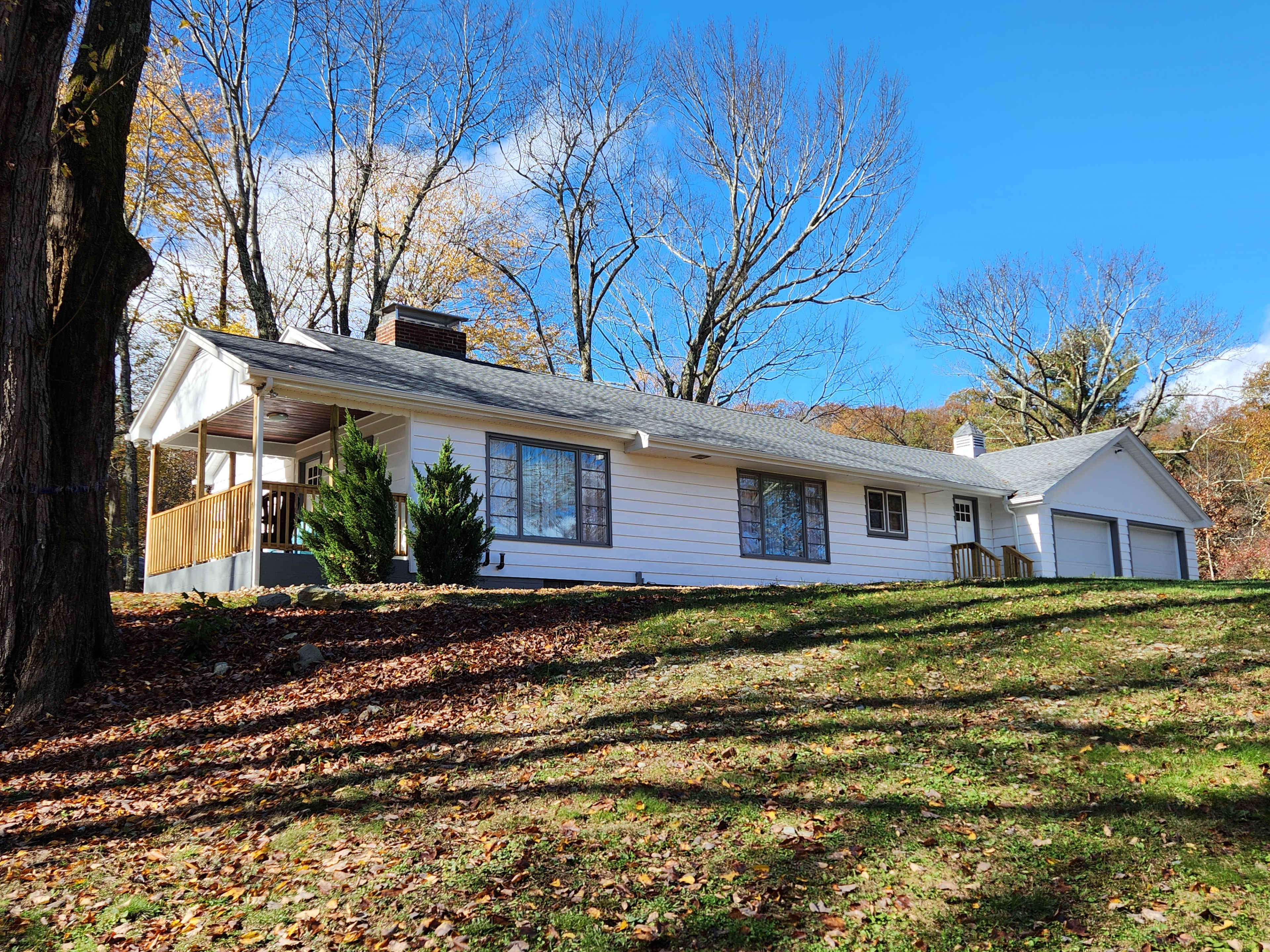 A single-story white house with a gray roof is situated on a hillside, surrounded by bare trees and autumn foliage.