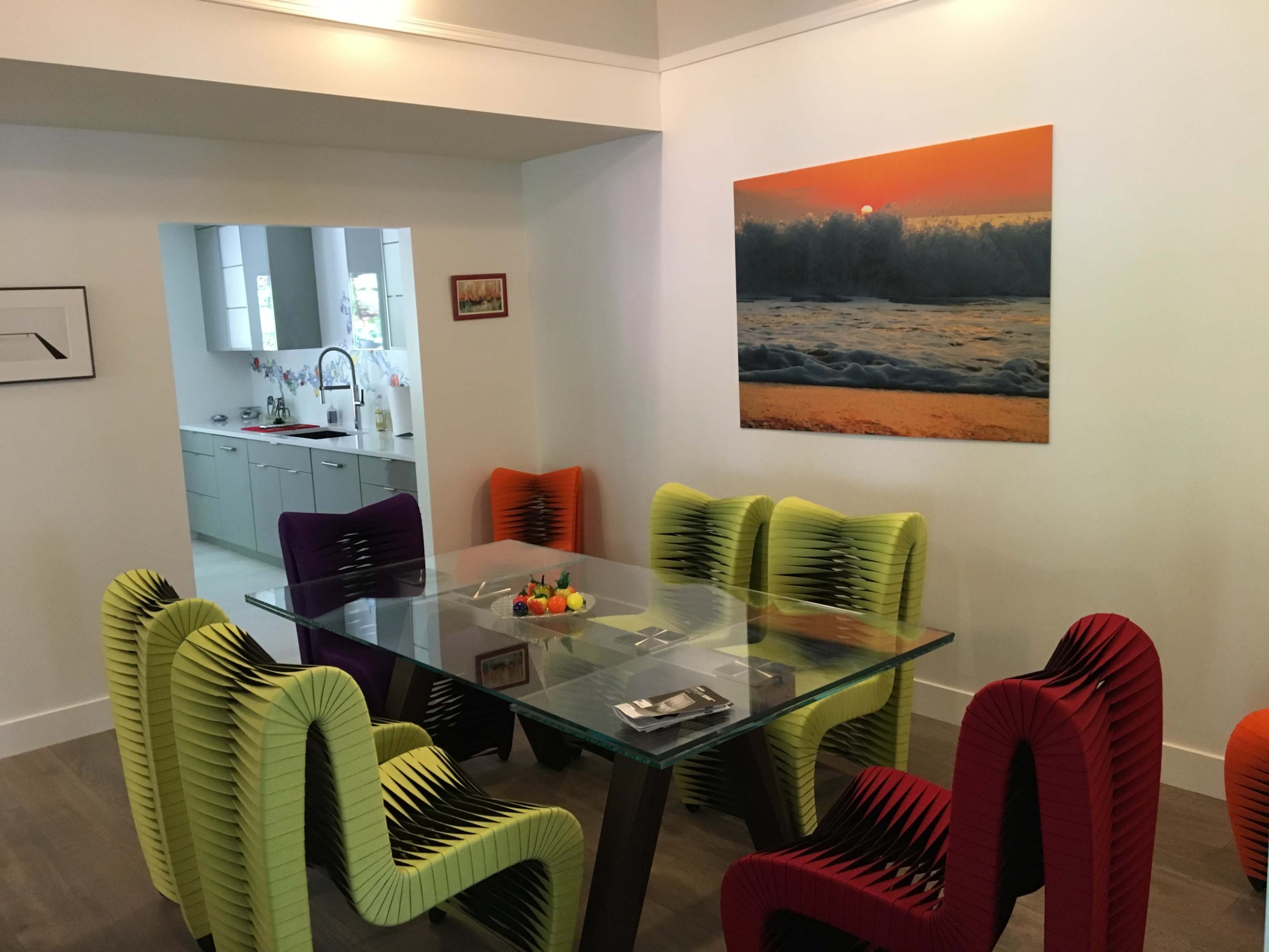 A modern dining area features a glass table surrounded by vibrant, uniquely designed chairs, with a large ocean-themed artwork on the wall.
