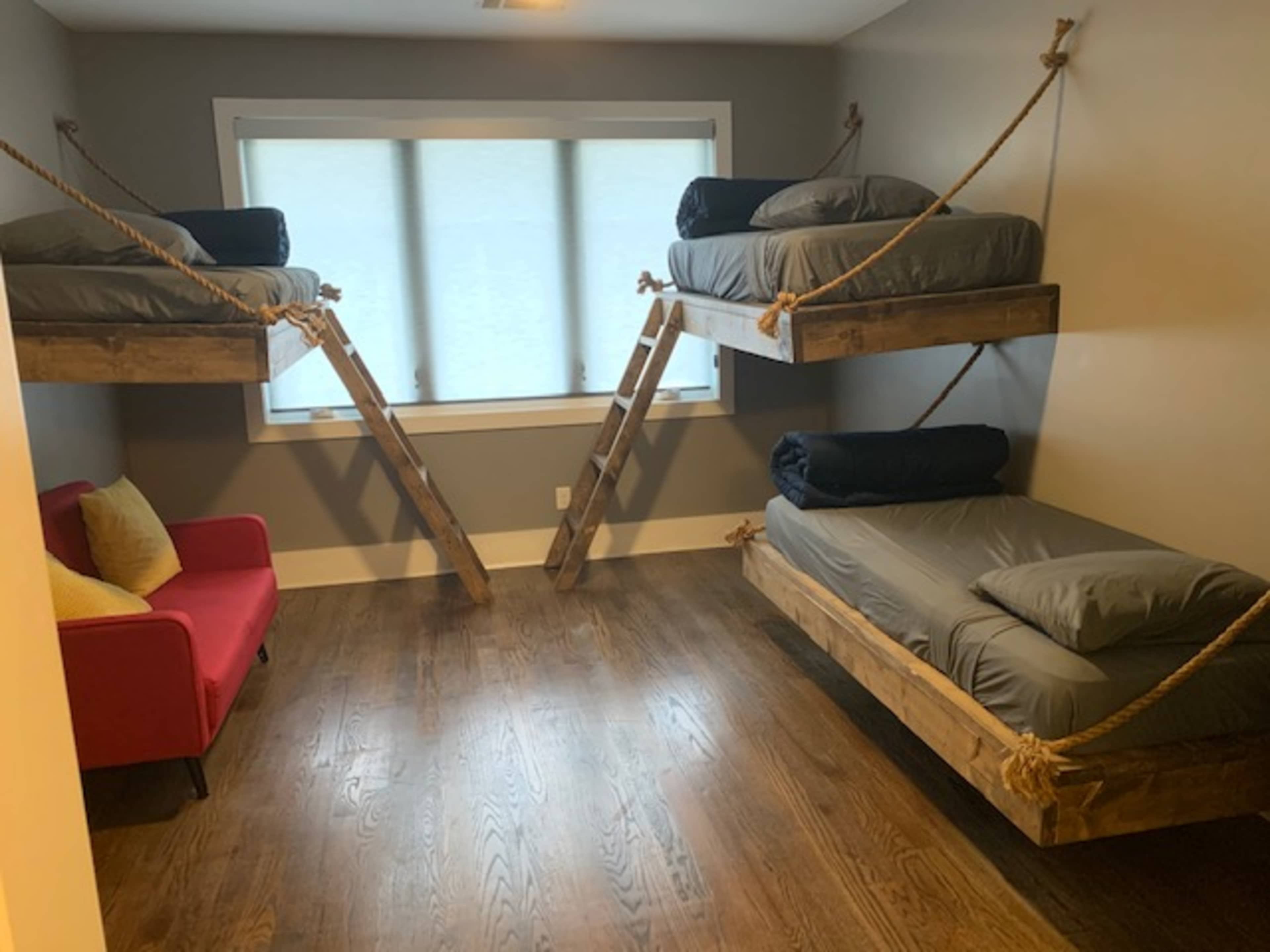 The image shows a room with two suspended bunk beds made of wood, with a red chair and large windows allowing natural light in.