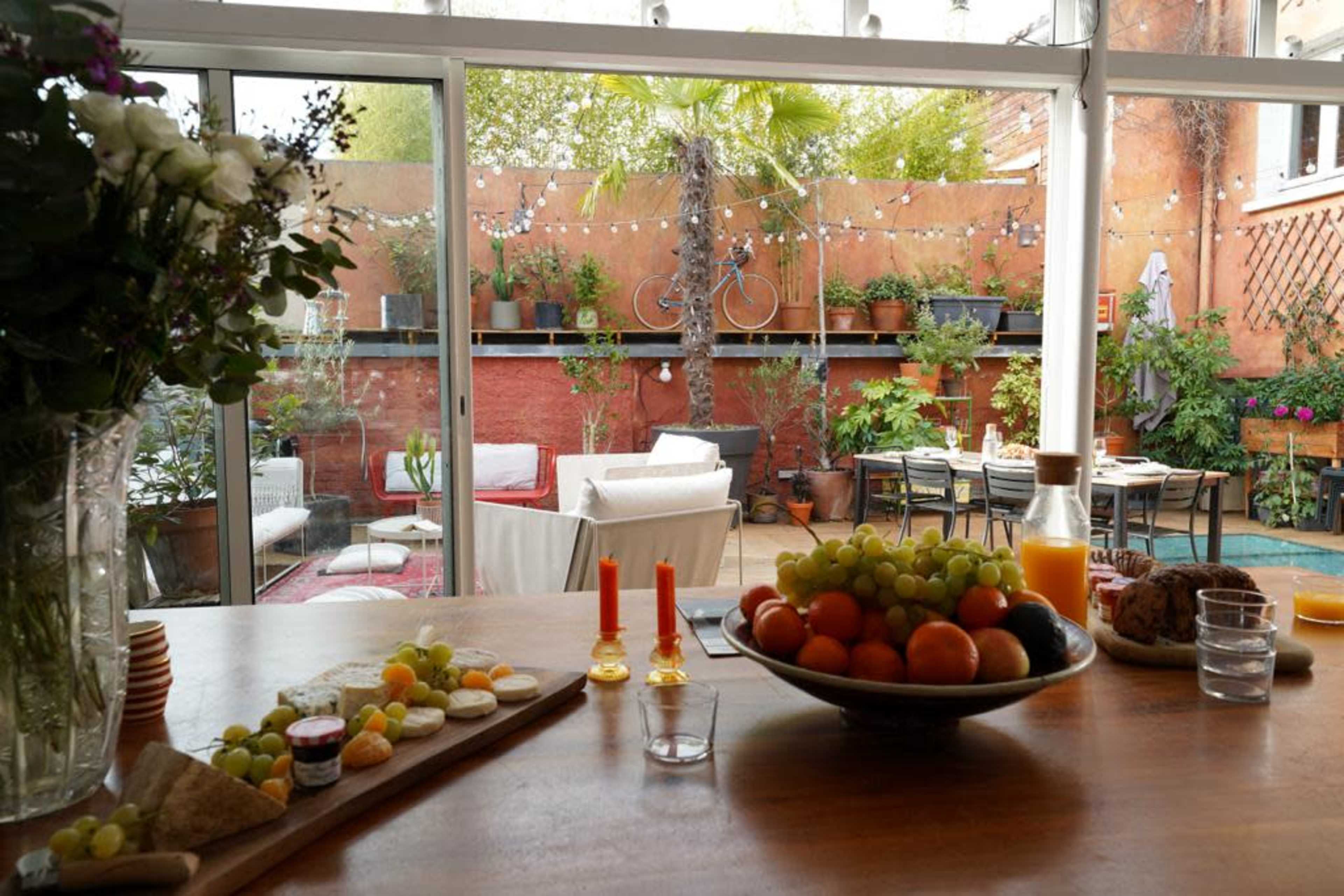A bright, indoor dining area features a wooden table set with fruits and snacks, overlooking a lush garden with outdoor seating and string lights.