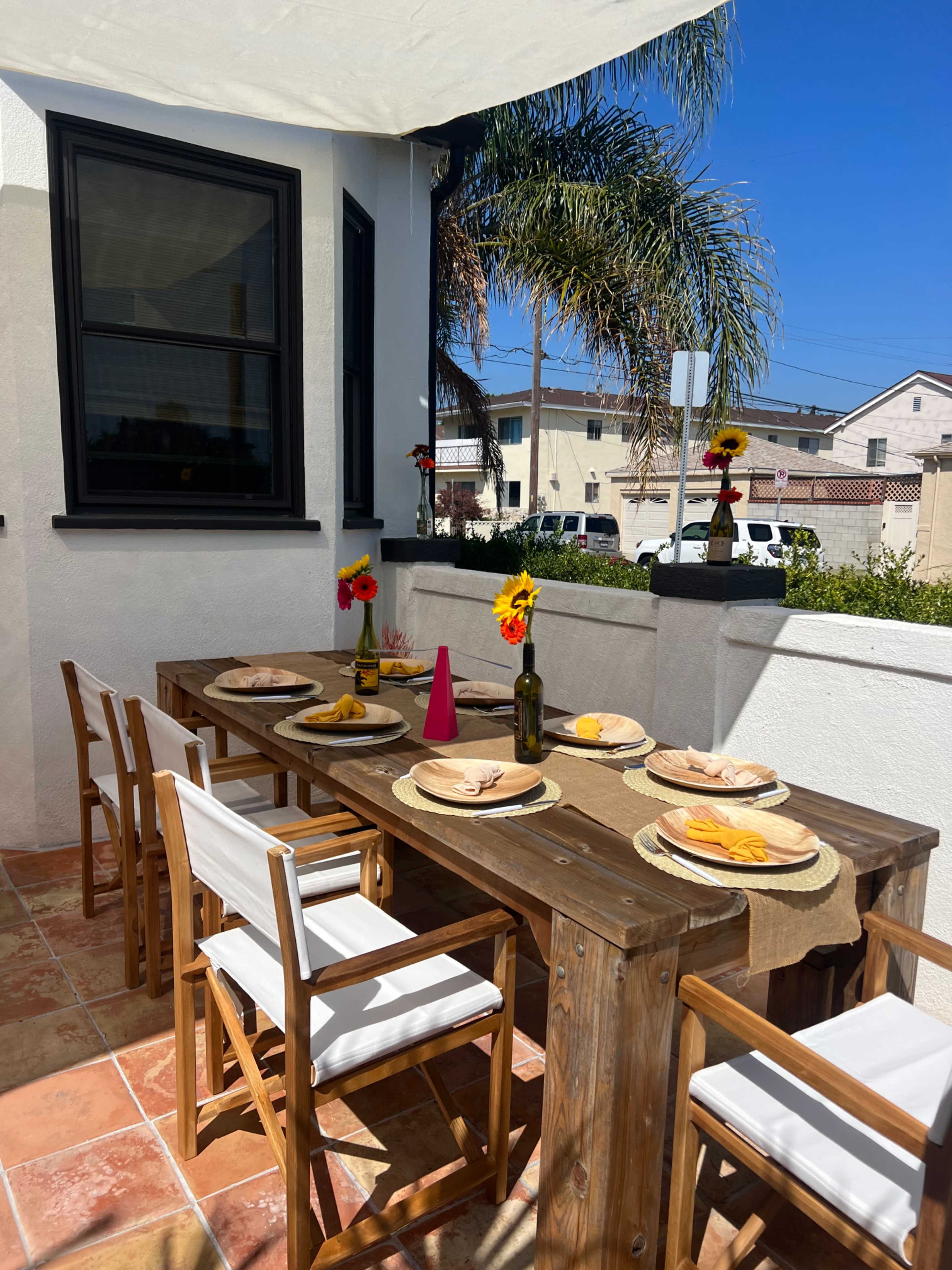 A wooden dining table is set for a meal on a patio, surrounded by chairs and decorated with flowers and dishes, with buildings and palm trees visible in the background.
