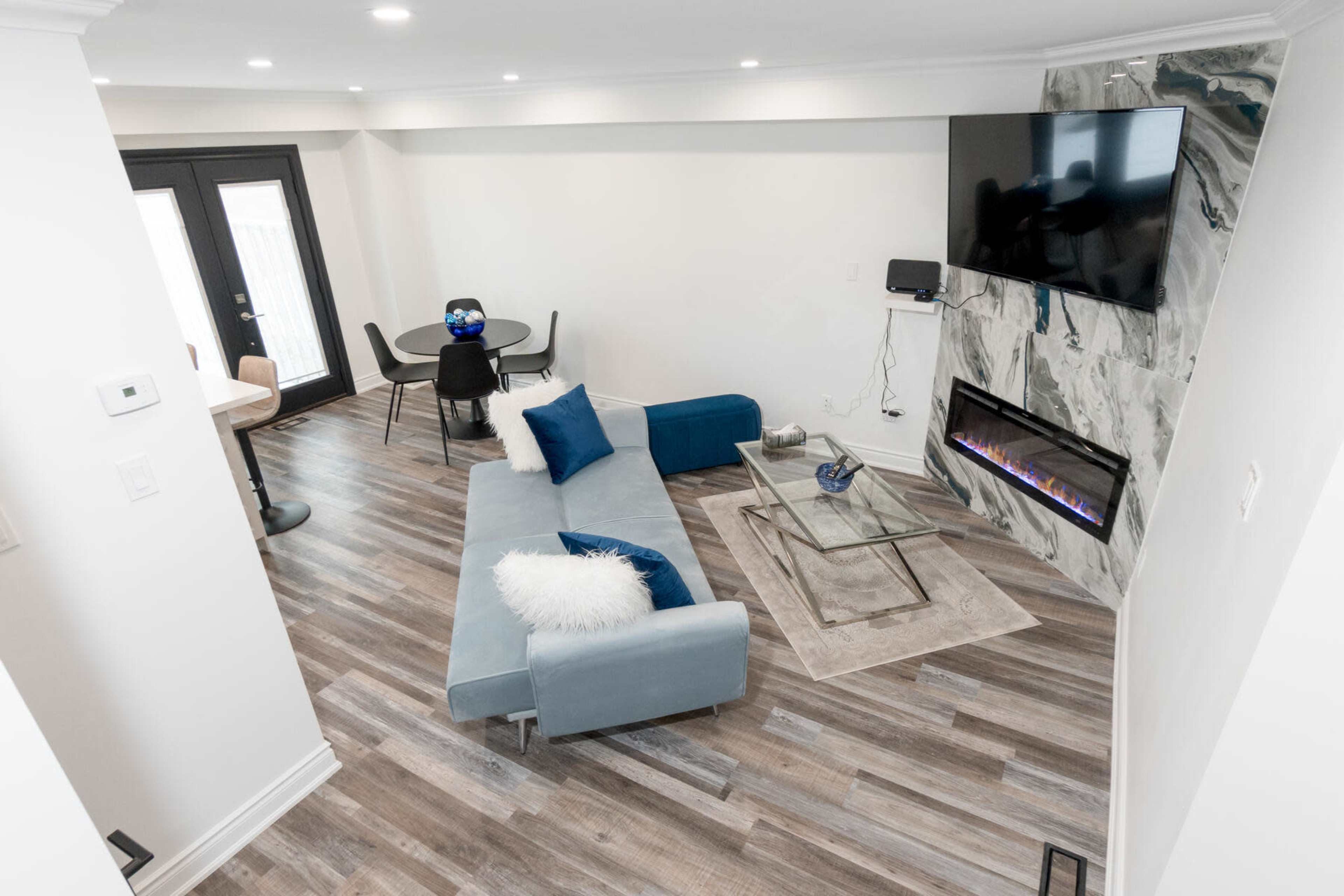 The image shows a modern living room with a blue sectional sofa, a glass coffee table, a wall-mounted TV, and a faux fireplace, all situated on a hardwood floor.