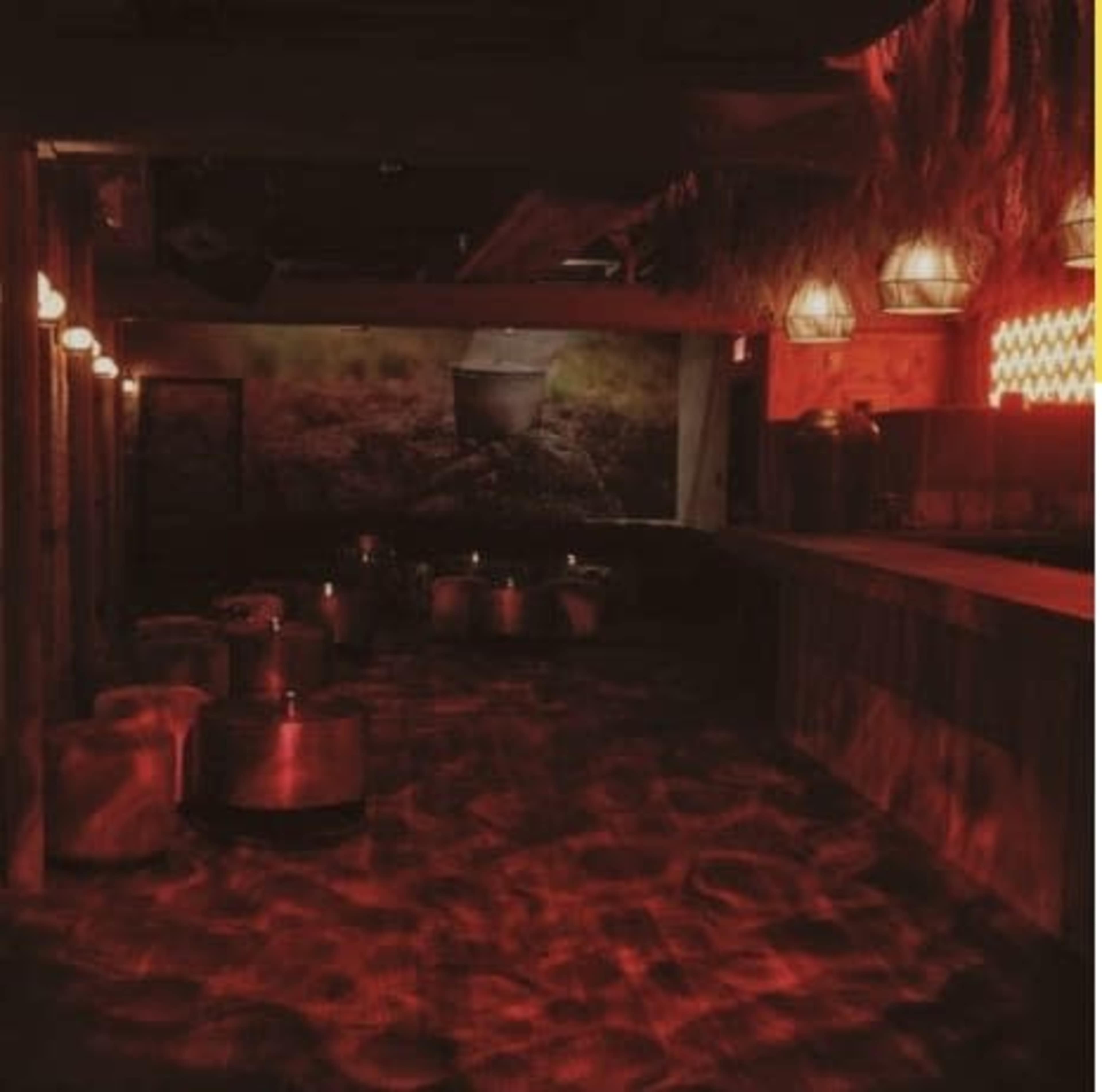 The image depicts a dimly lit room with seating arrangements made of cushions and metallic tables, illuminated by red light and adorned with tropical decor.