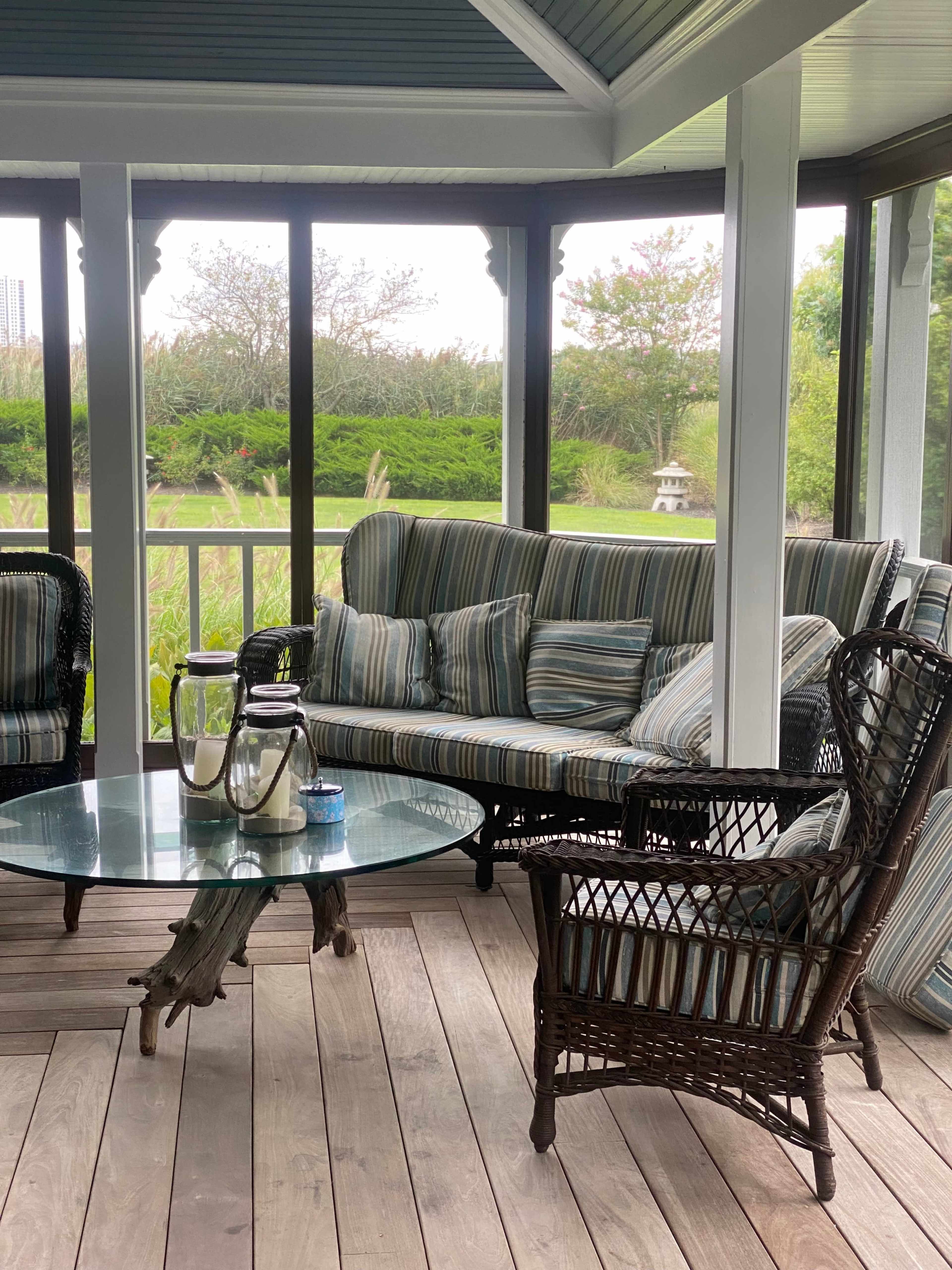 A screened porch with a striped sofa, wicker chairs, and a glass coffee table, overlooking a green landscape.