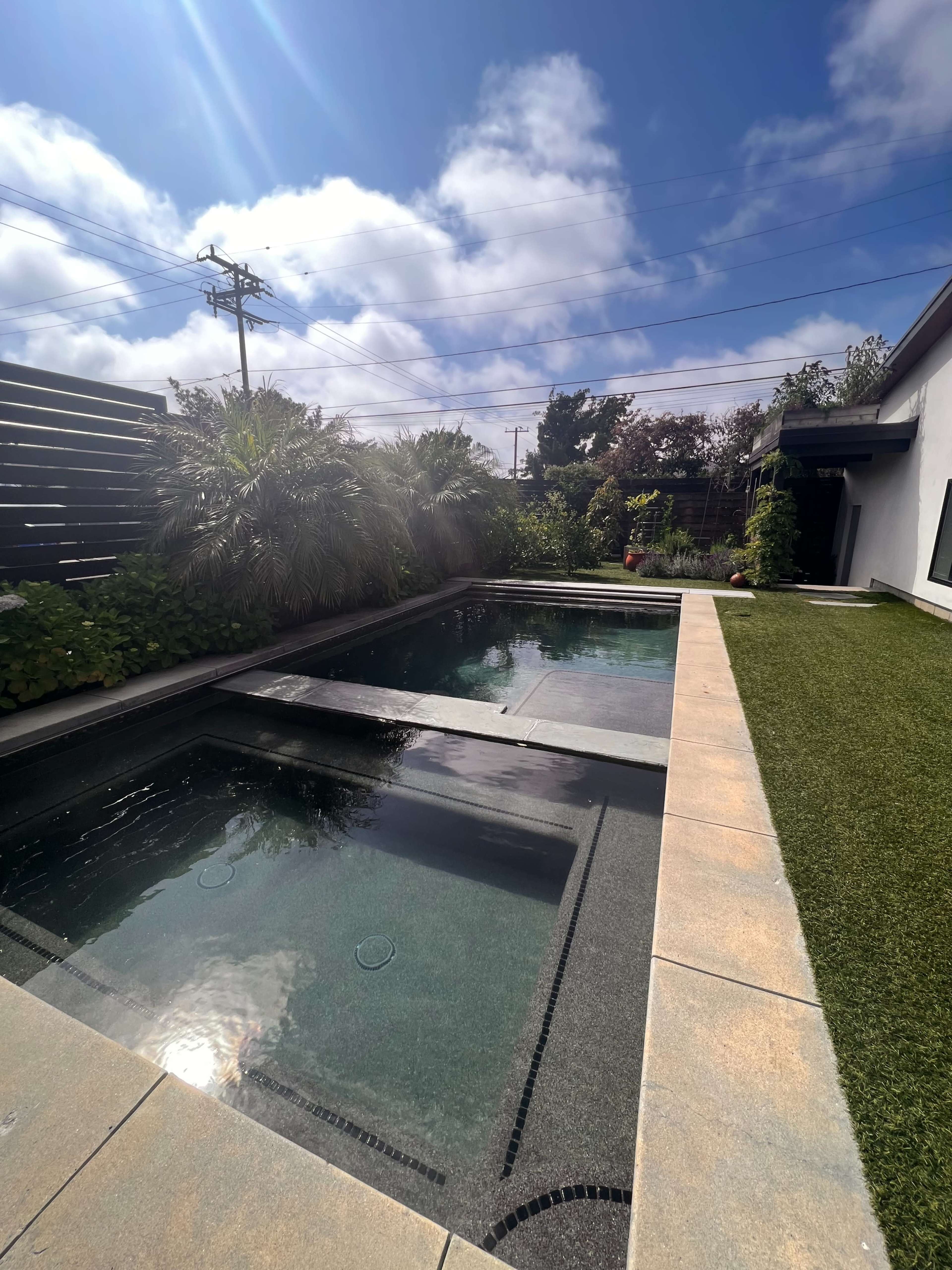 A modern outdoor space featuring two swimming pools surrounded by greenery and a patch of artificial grass under a partly cloudy sky.