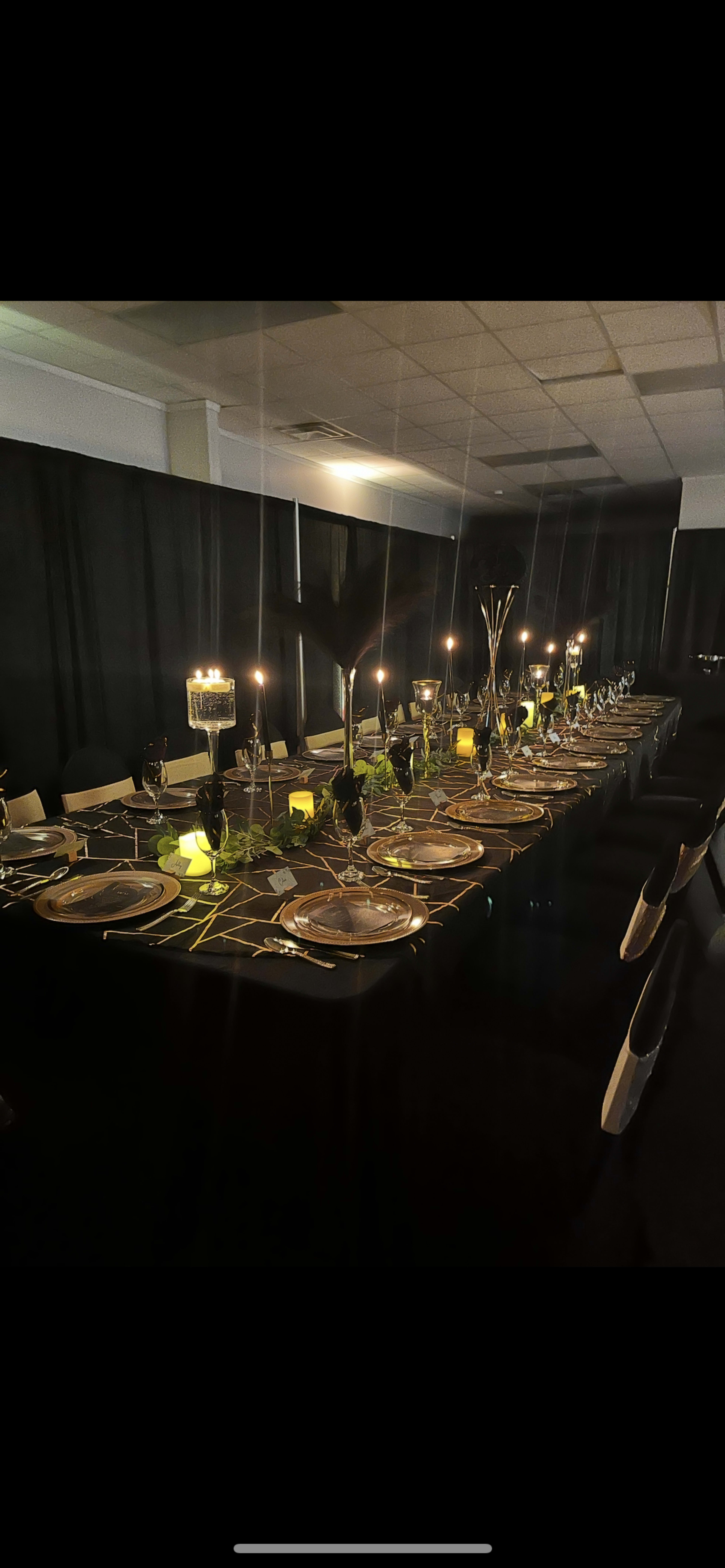 A long dining table is elegantly set with plates, glasses, and candles, surrounded by dark drapes and ambient lighting.
