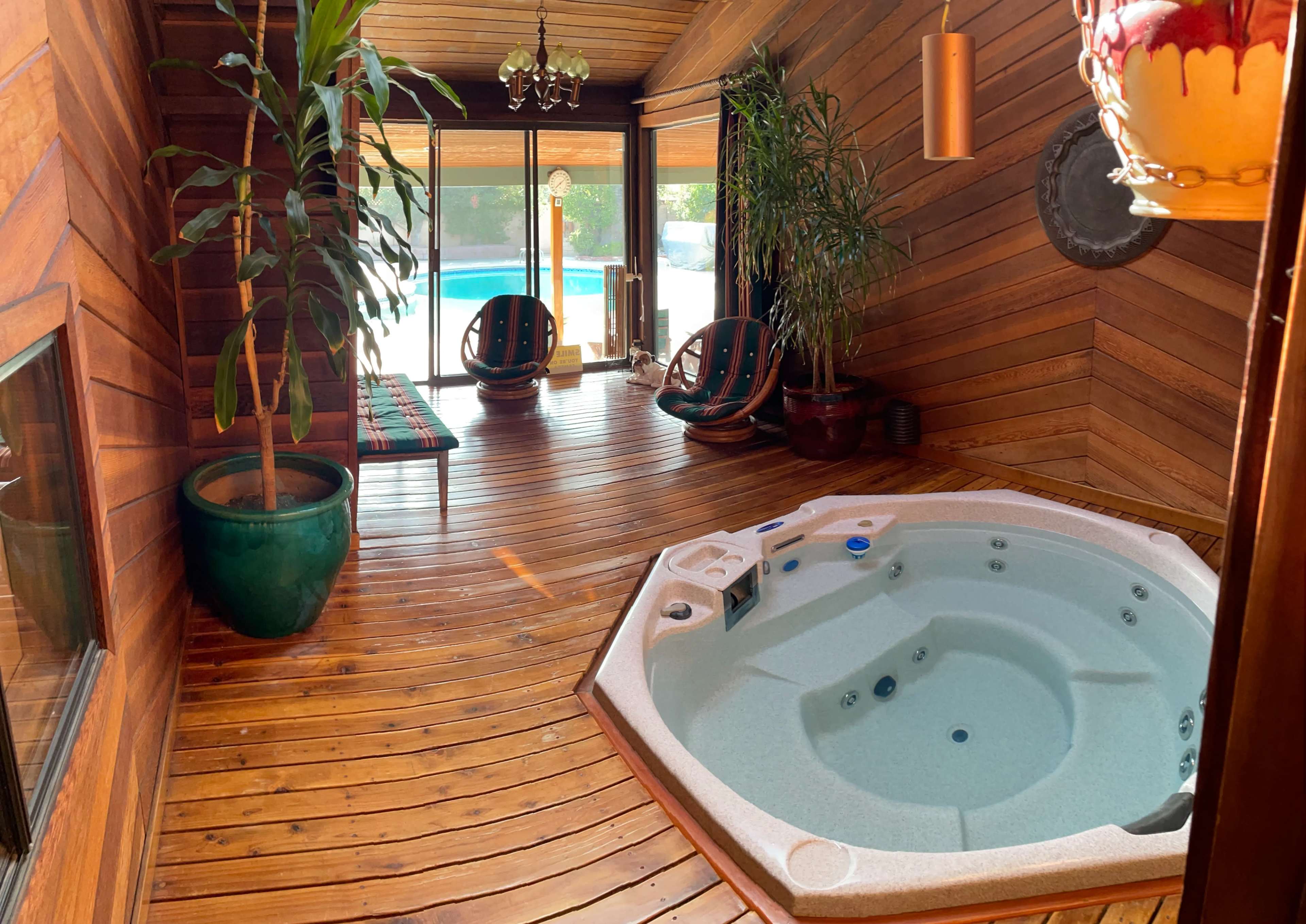 A wooden room featuring a hot tub, large windows, and two chairs beside potted plants.