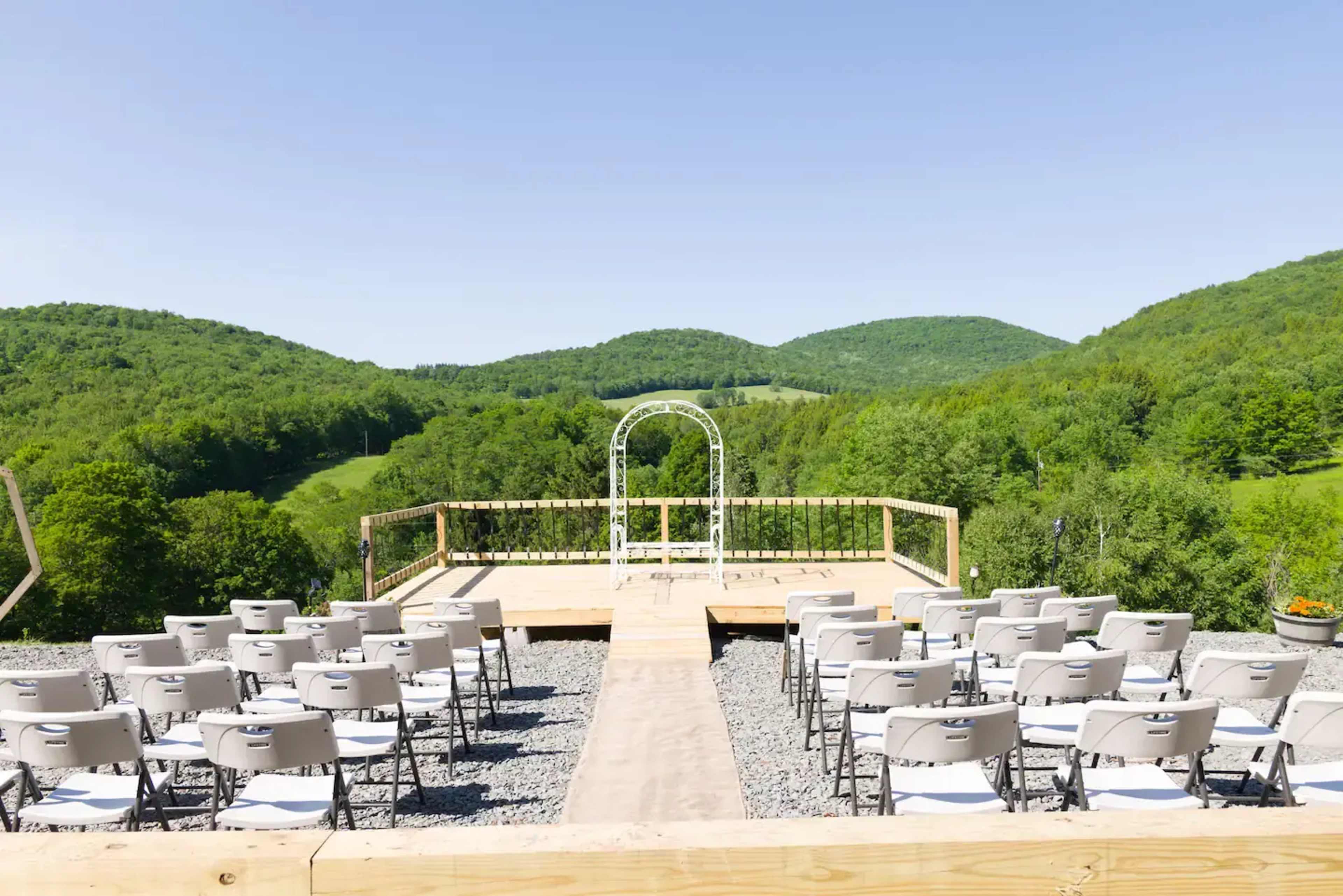 A wooden platform with white chairs arranged for an outdoor wedding ceremony overlooks green hills and a metal arch.