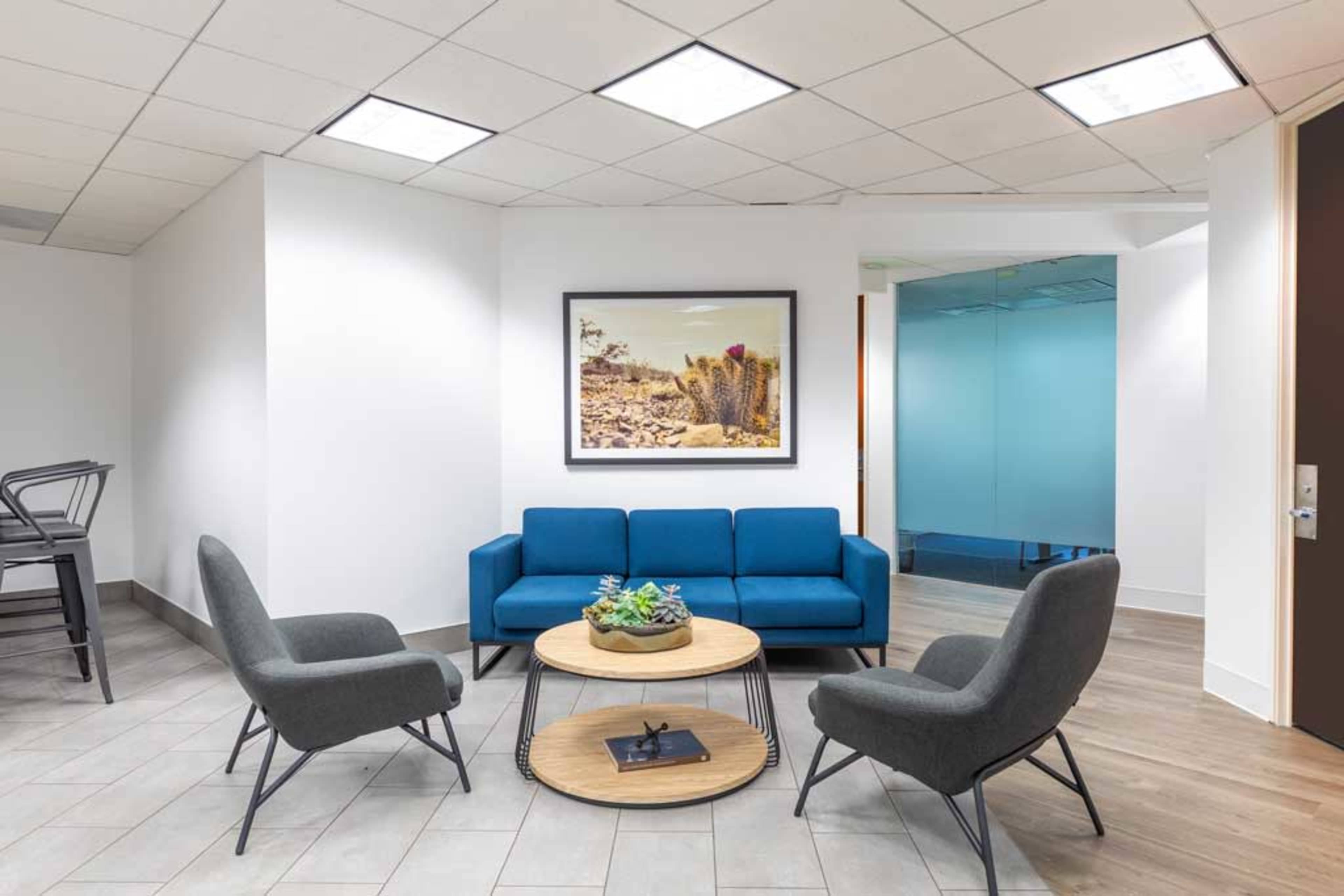 A modern office lounge area features a blue sofa, two gray armchairs, a round table with a plant centerpiece, and artwork on the wall.