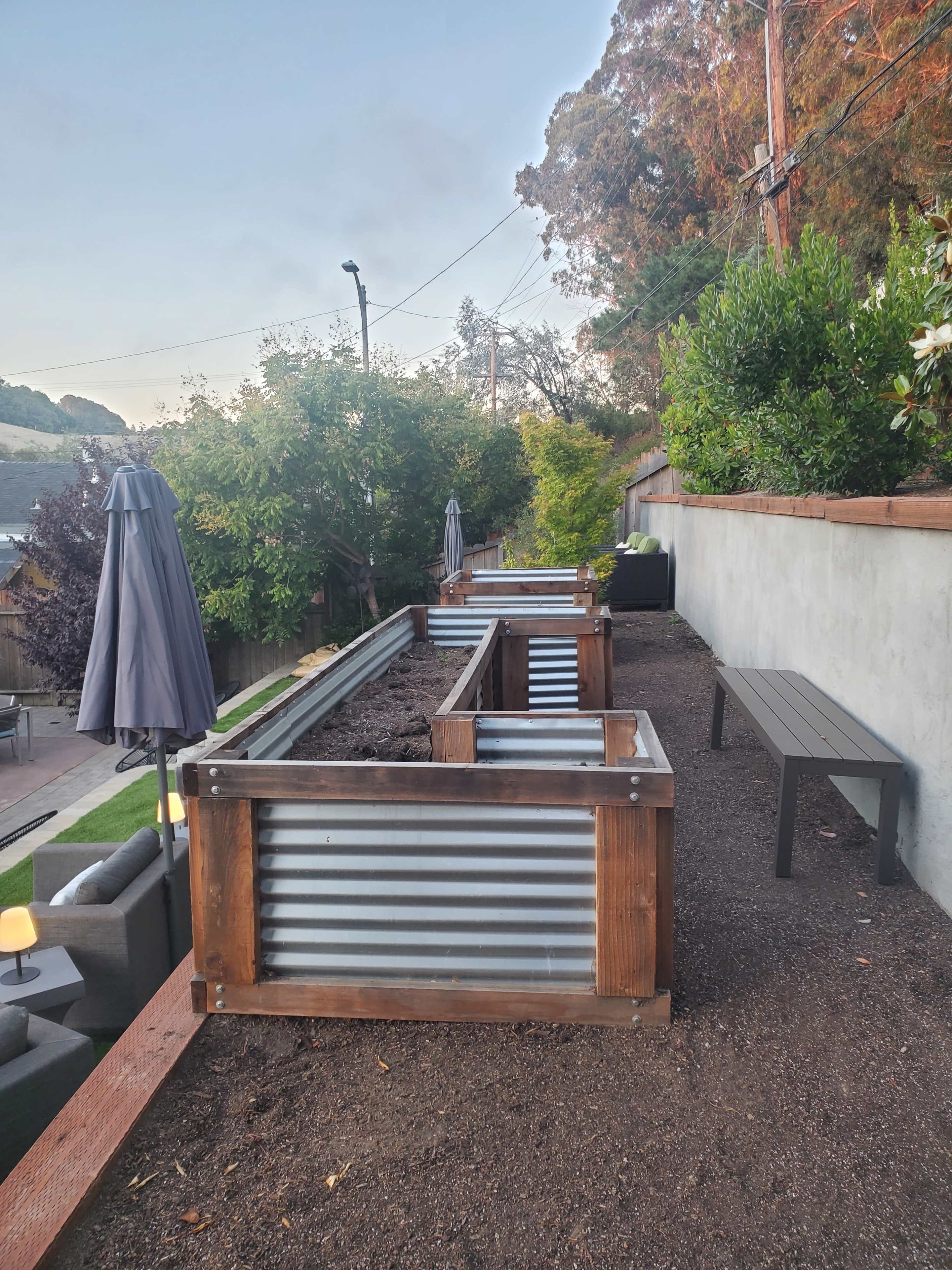 A landscaped area with metal garden beds, a wooden bench, and umbrellas situated on a hillside.