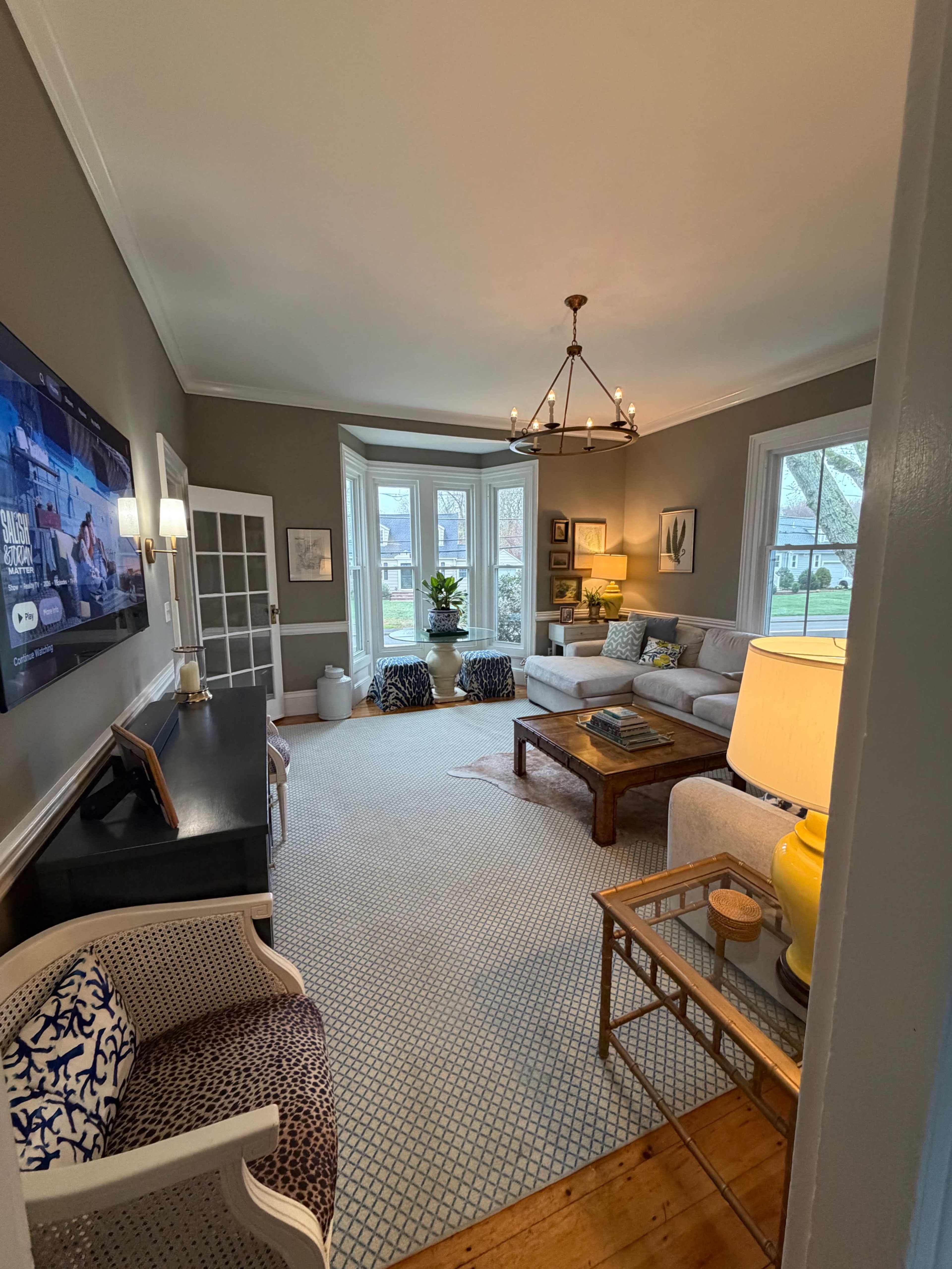 The image shows a cozy living room with a gray color scheme, a large window, a wooden coffee table, and seating arrangements that include a sofa and two patterned chairs.