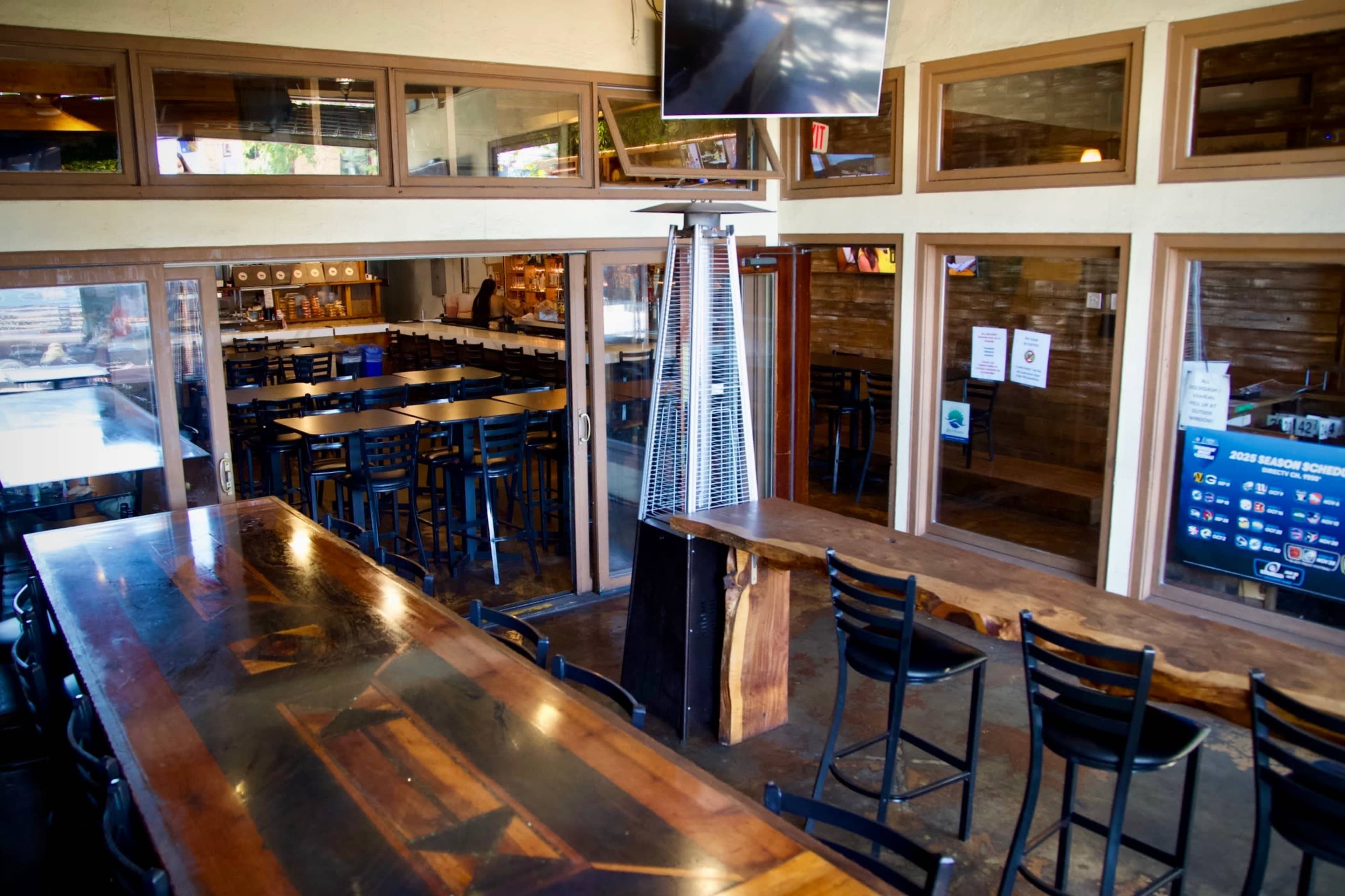 The image shows a spacious, well-lit dining area of a restaurant with a long wooden table, black chairs, and large windows.