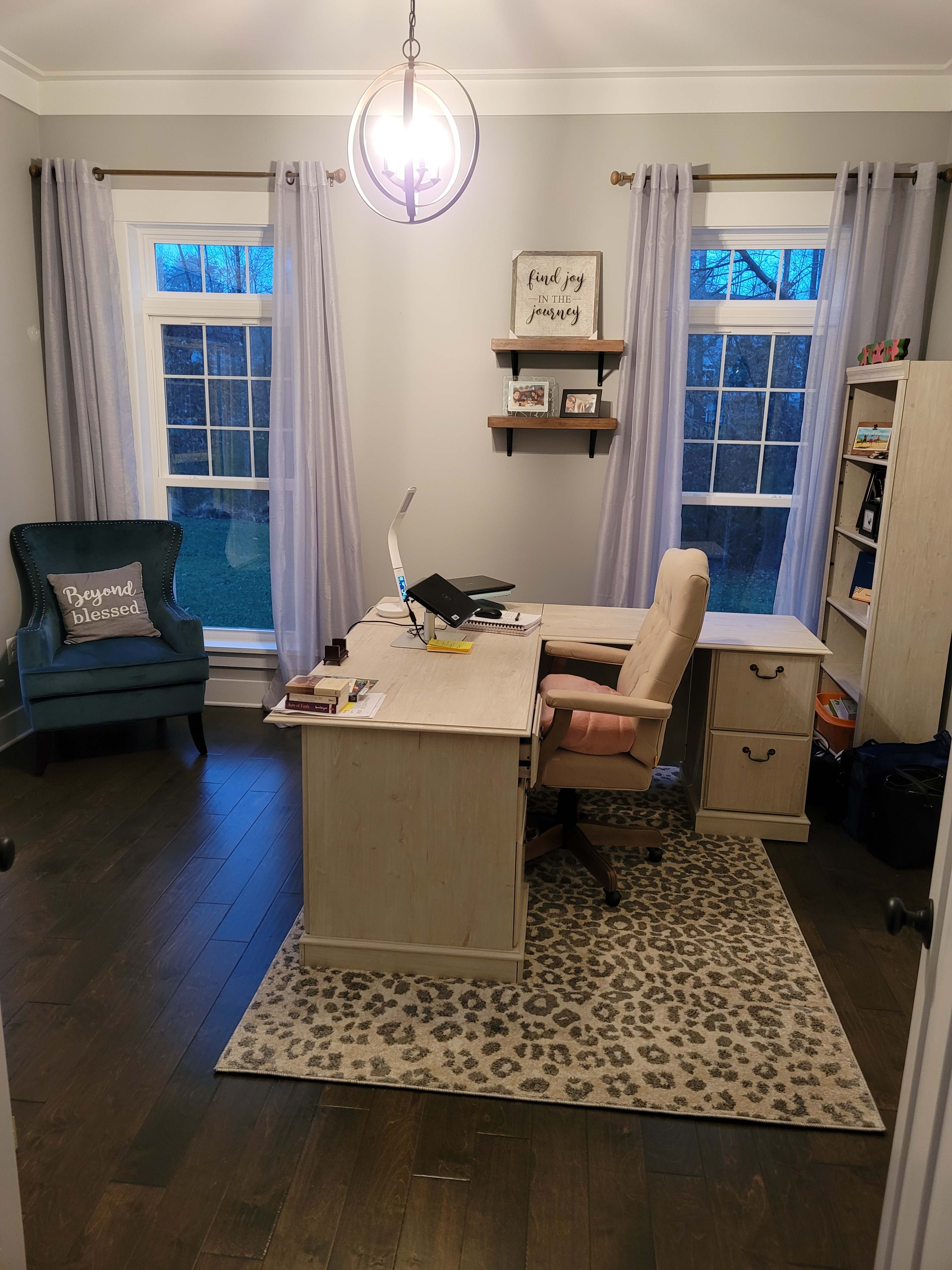 A home office with a desk, a chair, bookshelves, a large window, and a decorative rug.