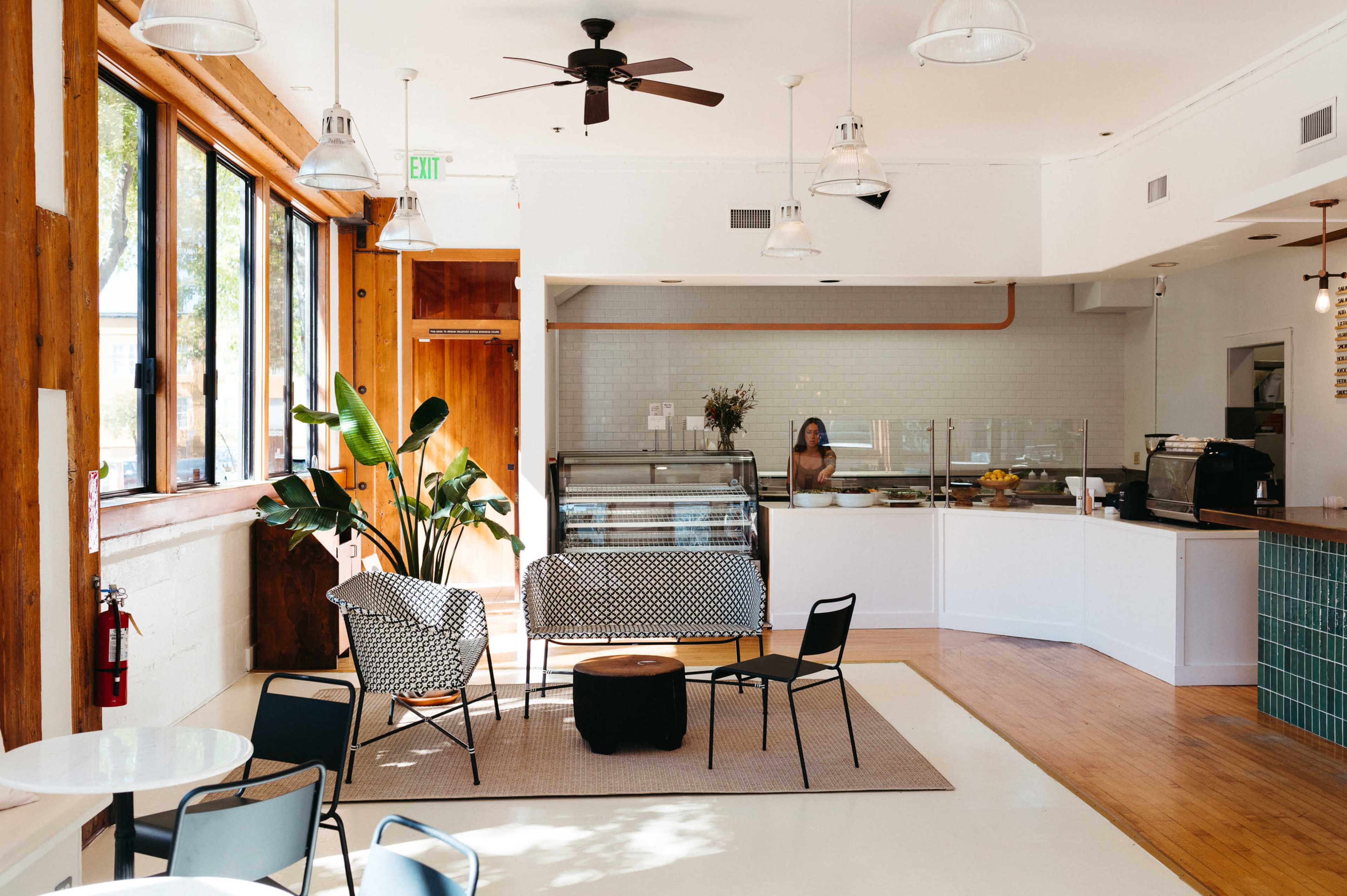 A spacious café features wooden accents, large windows, and a seating area with patterned chairs and a counter displaying food items.
